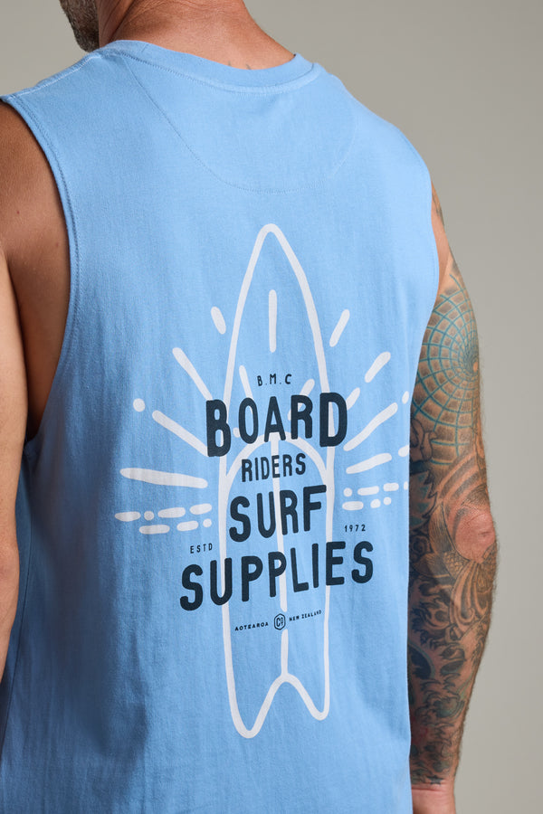 Wearing the Barkers BMC Board Riders Print Singlet in light blue, featuring a surfboard graphic and "Board Riders Surf Supplies" on the back, this organic cotton top is the perfect summer staple—especially paired with a tattooed right arm.