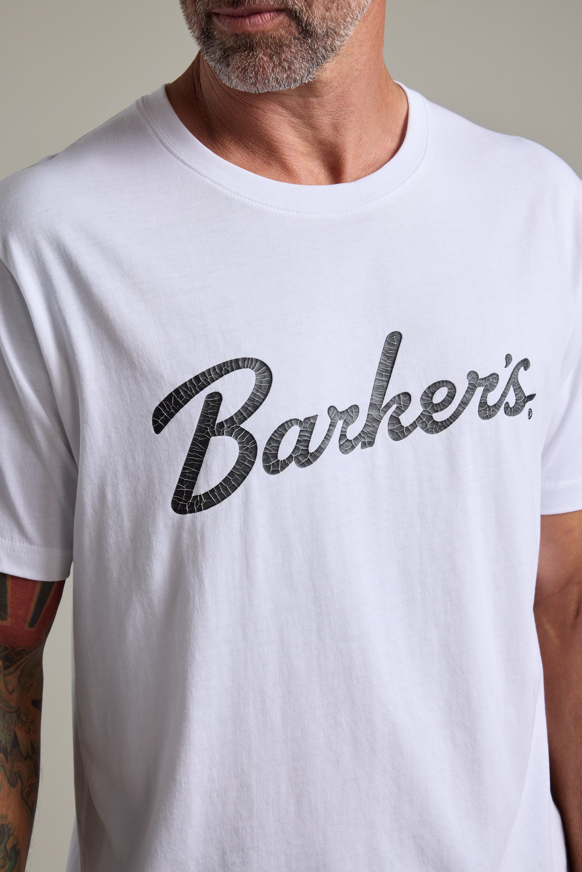 A man wears the Barkers Finely Tuned Print Tee, an organic cotton t-shirt with a vintage "Barkers" crackle print. Only the lower half of his face and upper torso are shown.