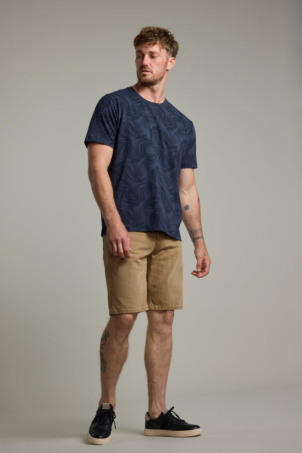 A man with short hair and tattoos poses against a plain background, wearing the Barkers Palm Leaves Print Tee in navy blue, tan shorts, and black sneakers. He stands relaxed, slightly looking to his left.