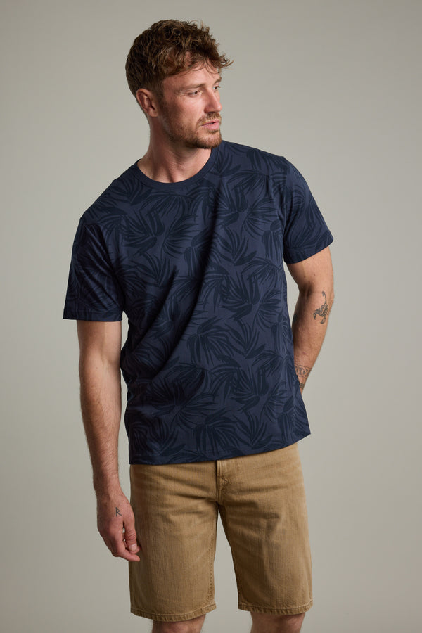 A man with short brown hair and a beard models the Barkers Palm Leaves Print Tee with tan shorts, standing against a light gray background and looking to his left, one arm at his side and the other slightly bent.