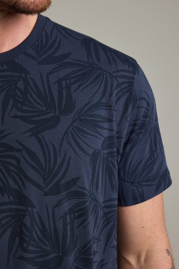 A close-up of a person wearing the Barkers Palm Leaves Print Tee, a regular fit navy blue organic cotton shirt with a dark palm print. Only the lower half of the face, shoulder, and upper arm are visible against a plain background.