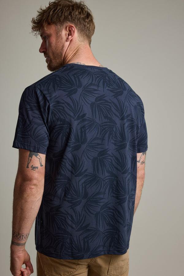 A man with tattooed arms, short light brown hair, and a beard wears the Barkers Palm Leaves Print Tee in navy blue and khaki pants. The plain background emphasizes his laid-back style.