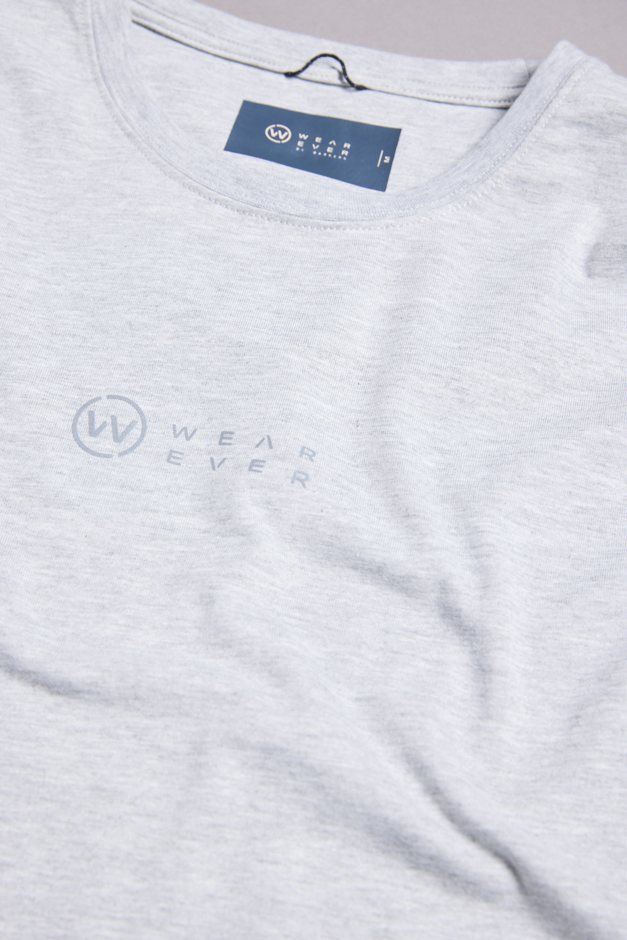 A close-up of the WearEver Outbound T-Shirt in light gray, featuring the subtle “Wear Ever” logo and text on the chest. The neckline displays a visible WearEver brand label.