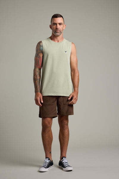 A bearded man with short hair stands facing forward, wearing the Barkers Newton Washed Singlet in light green, brown shorts, and black sneakers. A colorful tattoo sleeve covers his right arm. The plain backdrop adds a vintage feel.
