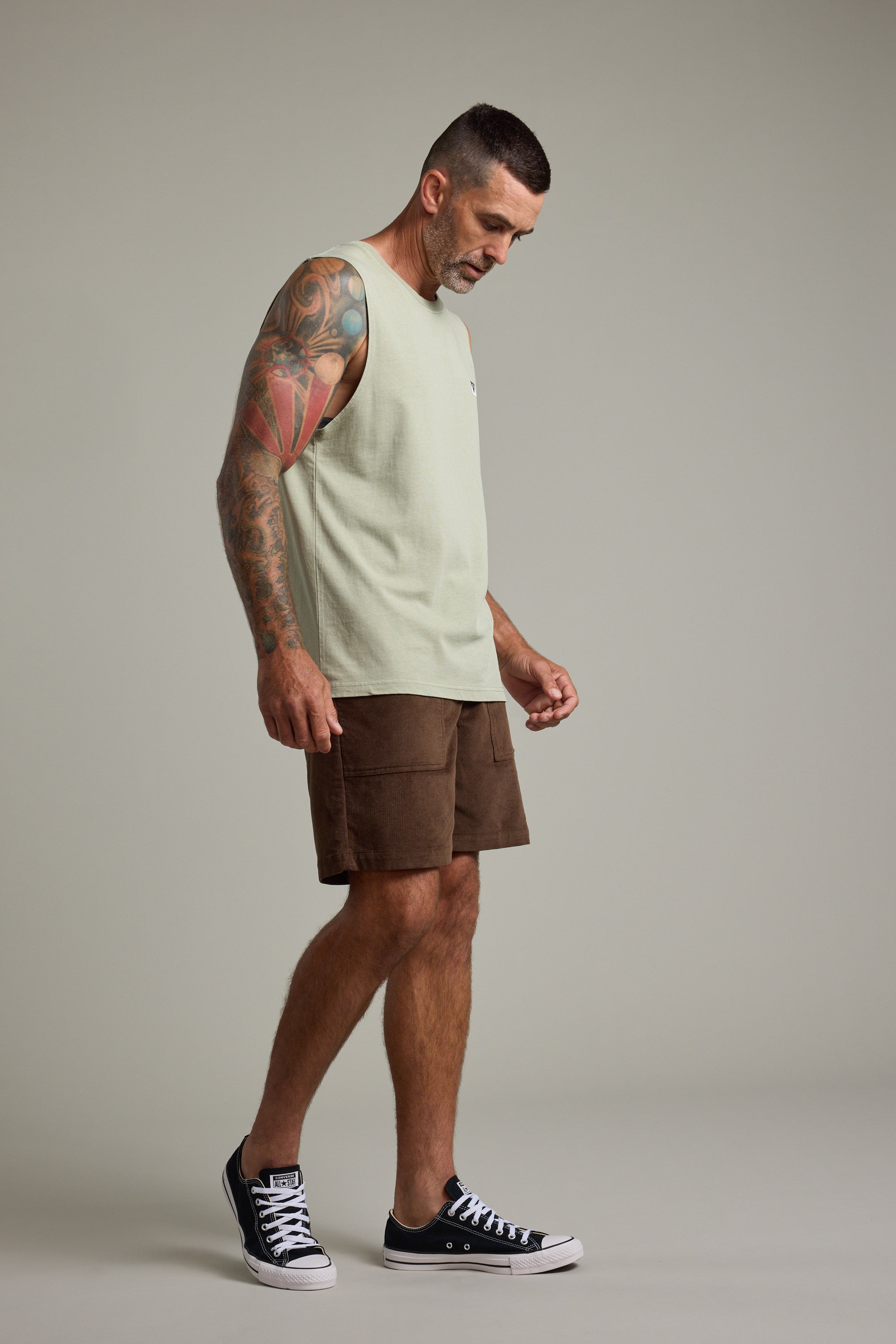 A man with short hair and tattooed arms wears the Barkers Newton Washed Singlet, styled with brown shorts and black sneakers. He stands against a plain background, looking down with a neutral expression.