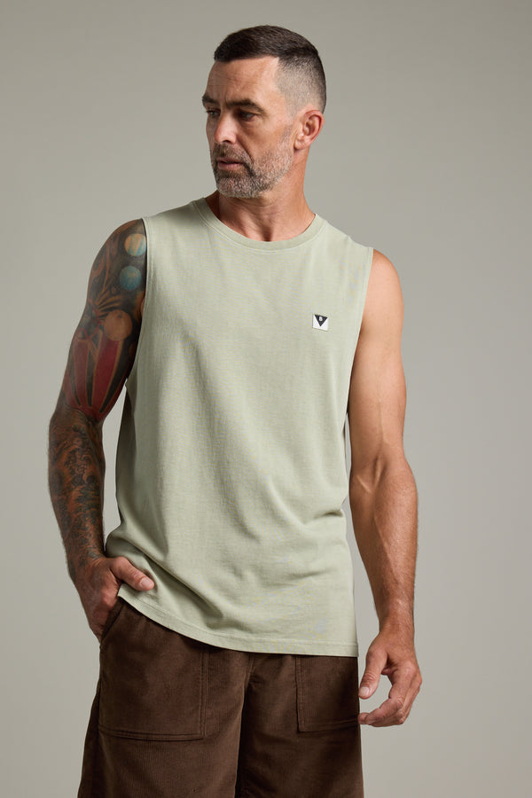 A man with short dark hair and a beard wears the Barkers Newton Washed Singlet in light green and brown shorts. With a colorful tattoo sleeve on his left arm, he looks left with one hand in his pocket, giving off a vintage vibe.