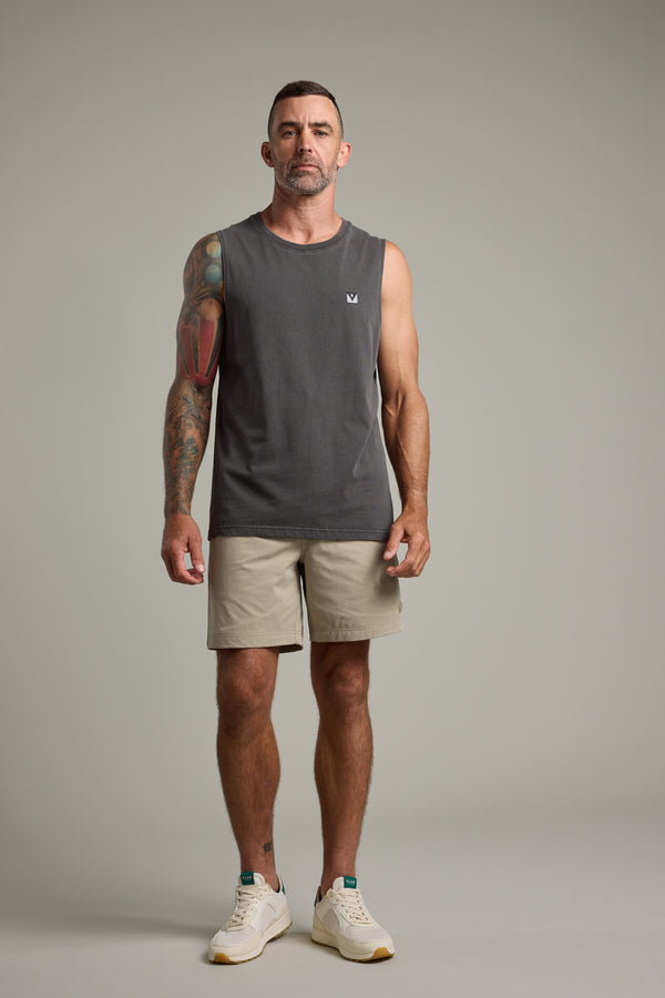 A man with short dark hair and a trimmed beard faces forward in a Barkers Newton Washed Singlet, dark gray, paired with beige shorts and white sneakers. He has colorful right-arm tattoos, a neutral expression, and stands against a plain background.