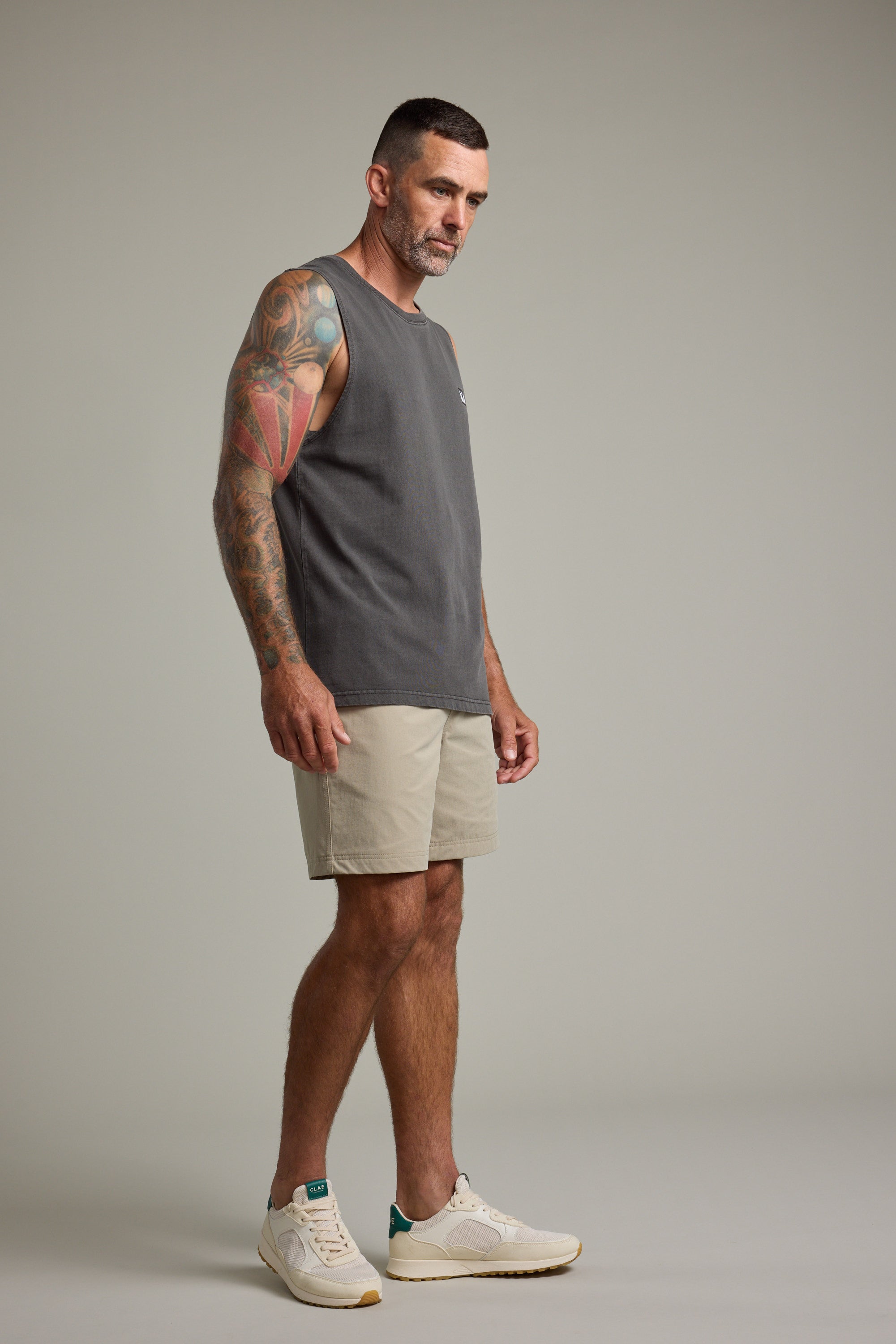A man with short dark hair and a tattooed arm wears the Barkers Newton Washed Singlet, paired with beige shorts and white sneakers. He stands in a neutral pose against a plain light gray background, creating a subtle vintage vibe.
