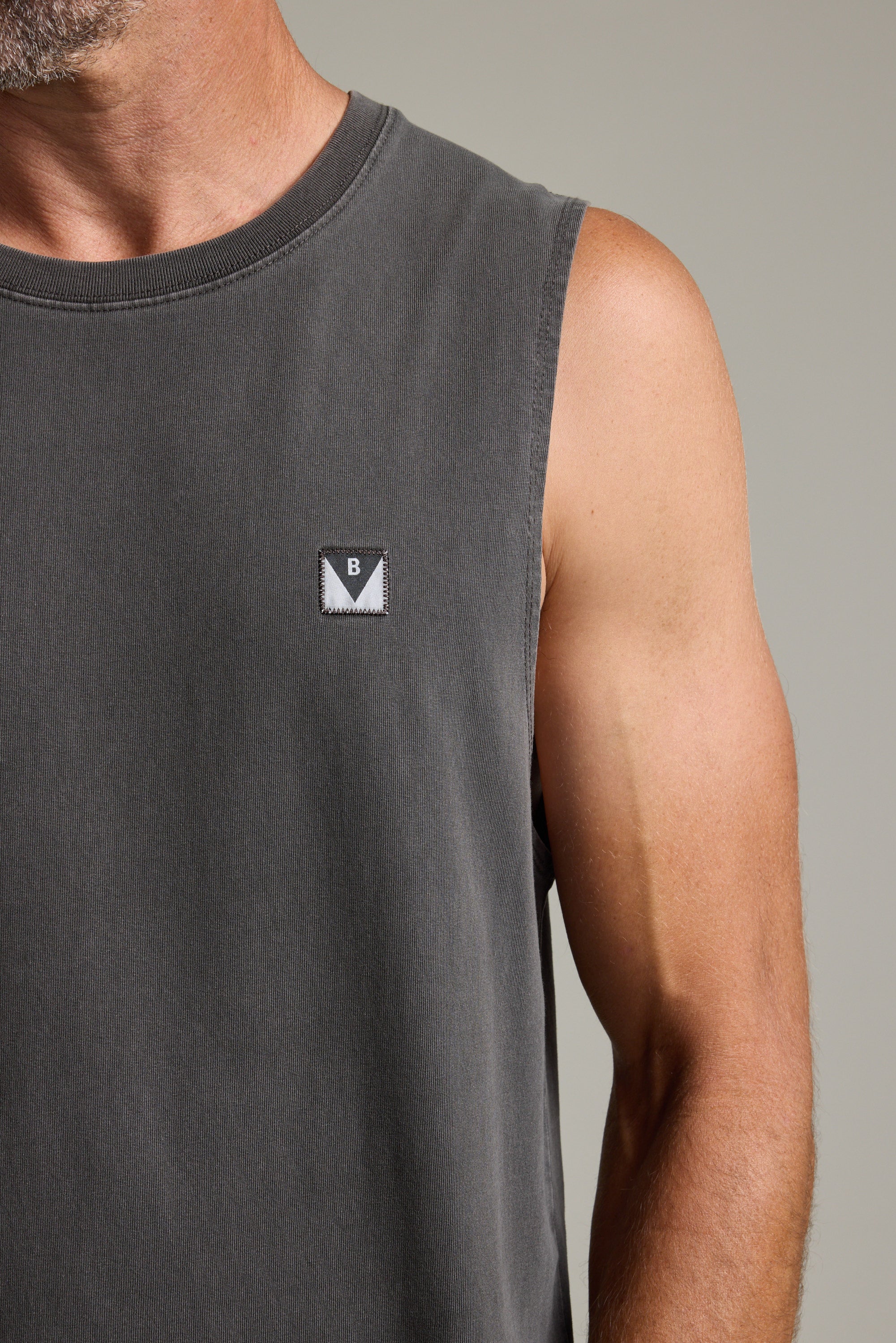 A close-up shows a person wearing the Barkers Newton Washed Singlet, an oversized sleeveless dark gray top with a small rectangular B patch on the chest. Only their lower face, shoulder, and upper arm are visible.