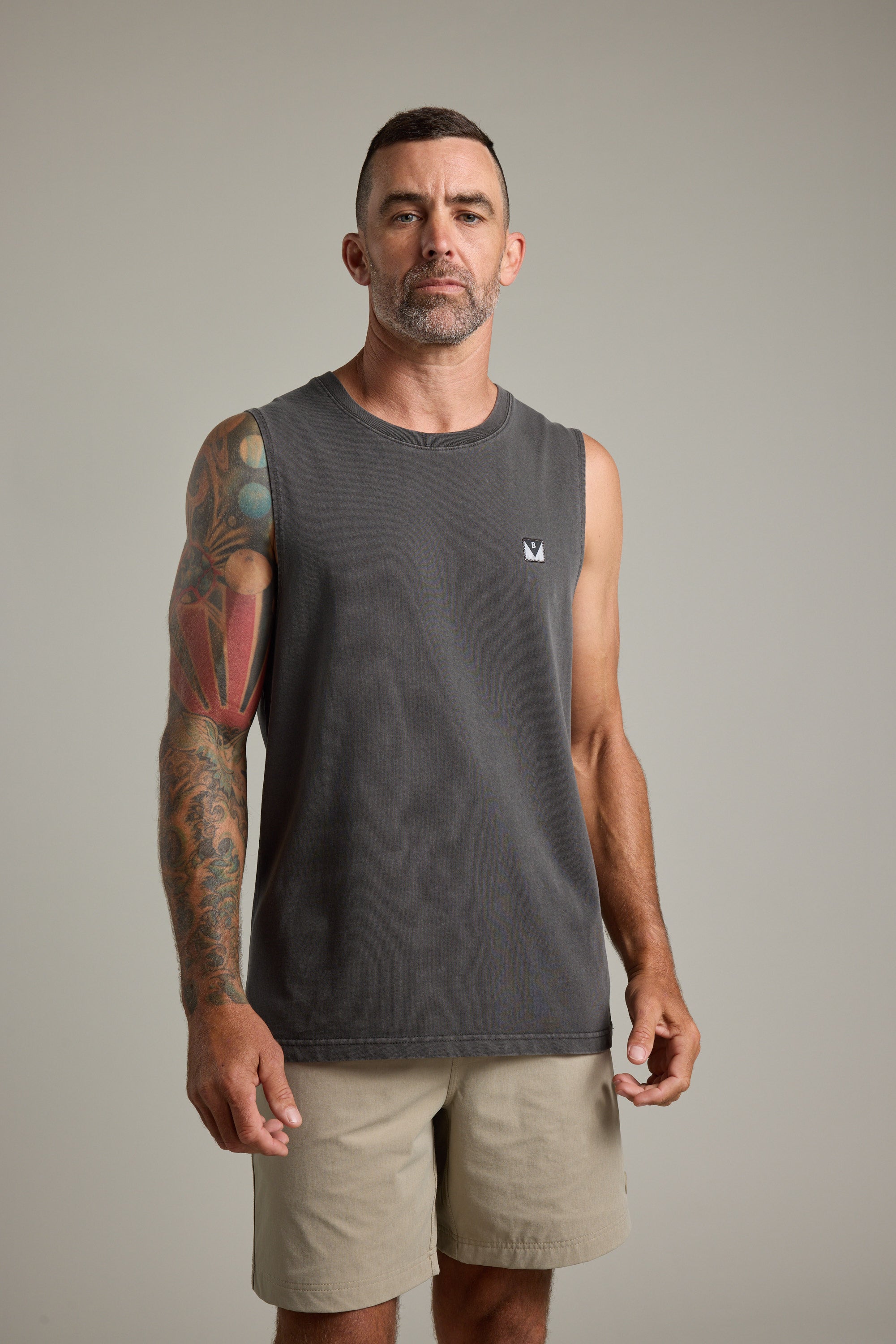 A man with short hair and a beard stands facing forward, wearing the Barkers Newton Washed Singlet in dark gray and light khaki shorts. His left arm's colorful tattoos stand out against the plain light gray background.