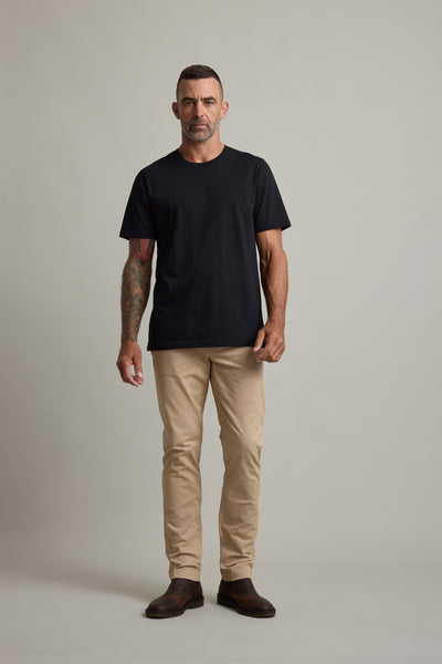Man in Barkers Good Earth Cotton Essential Tee, beige pants, and brown boots stands against backdrop.