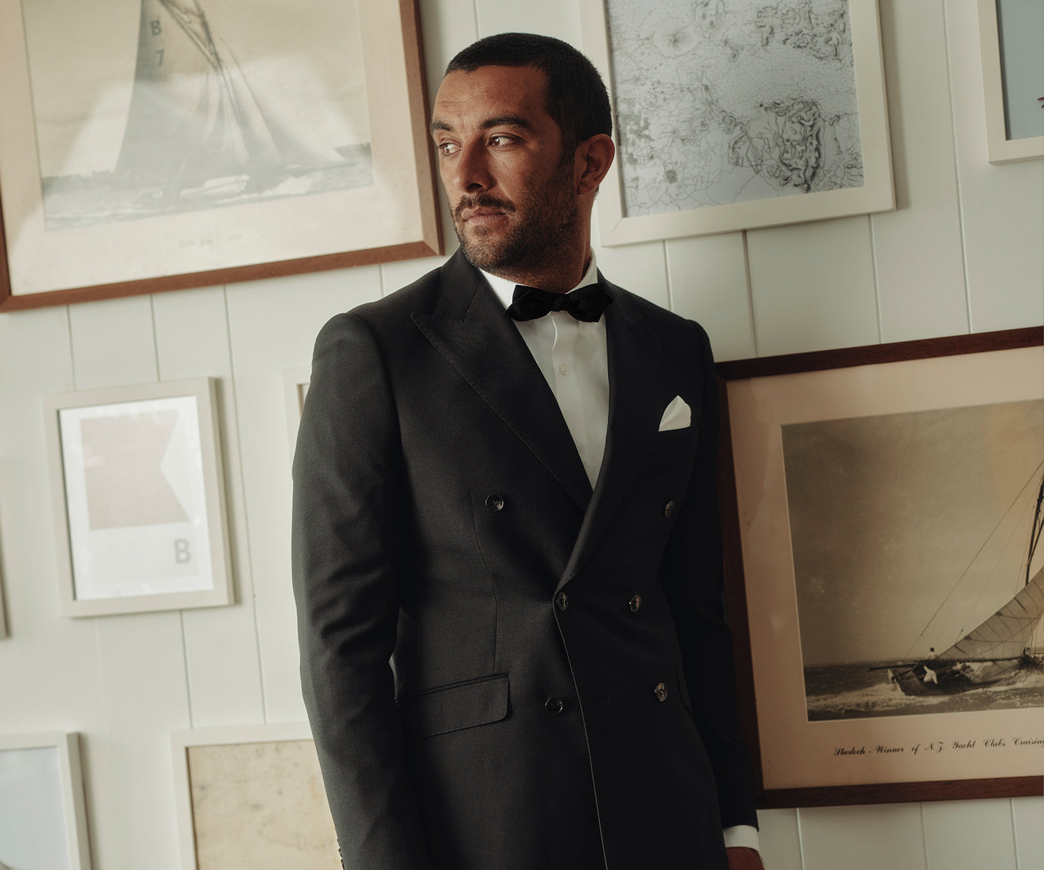 A man in a black tuxedo with a bow tie and white pocket square stands indoors, looking to the side. He is in front of a wall decorated with framed photographs and artwork.