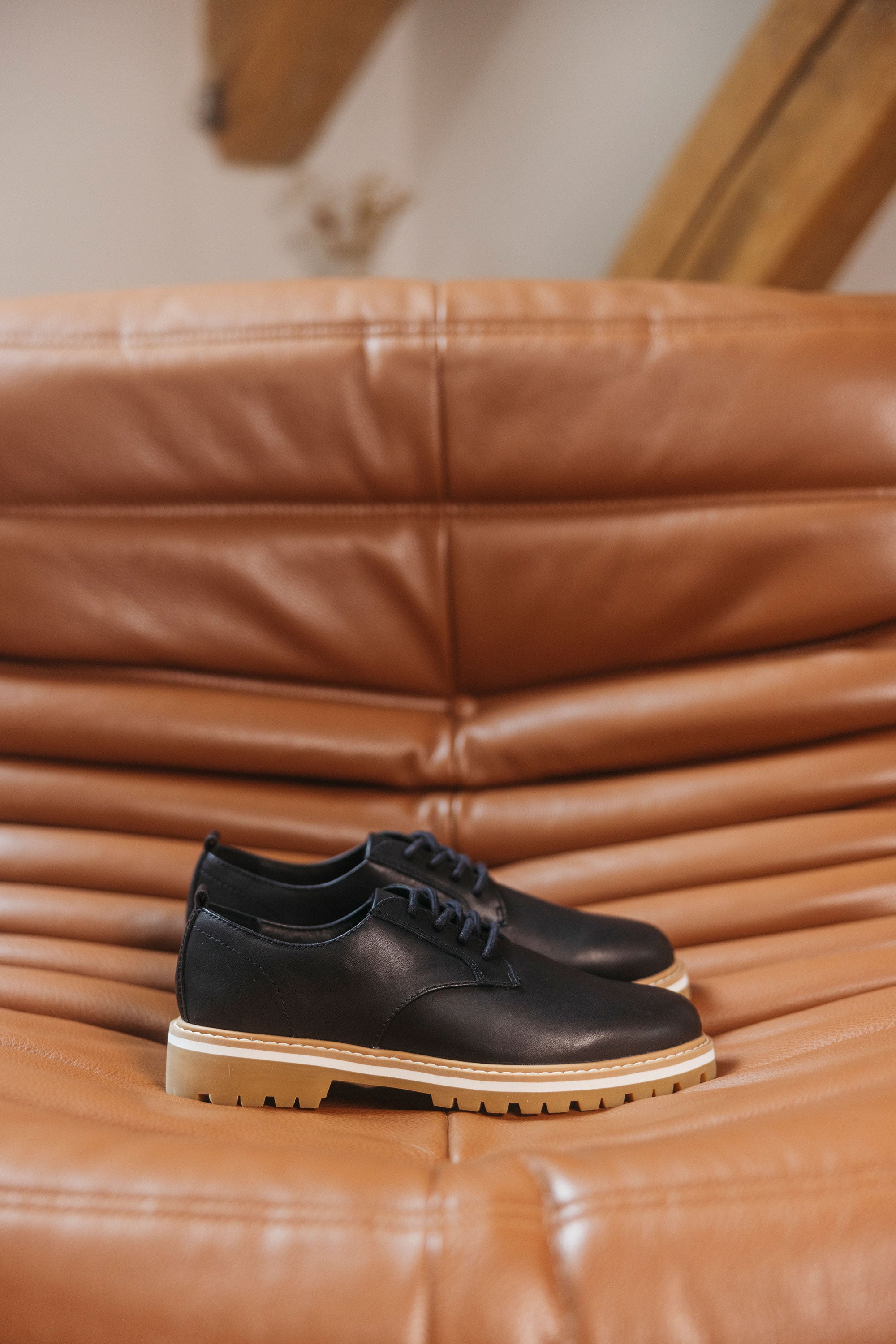 The CLAE Logan by Clae, black leather derby sneakers with light tan soles and recycled PU foam insoles, are displayed on a tan tufted leather couch.