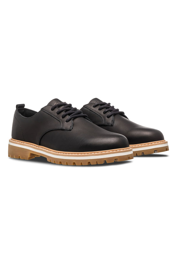 The CLAE Logan by Clae is a pair of black leather derby sneakers with light brown rugged rubber soles and a recycled PU foam insole, shown against a white background.