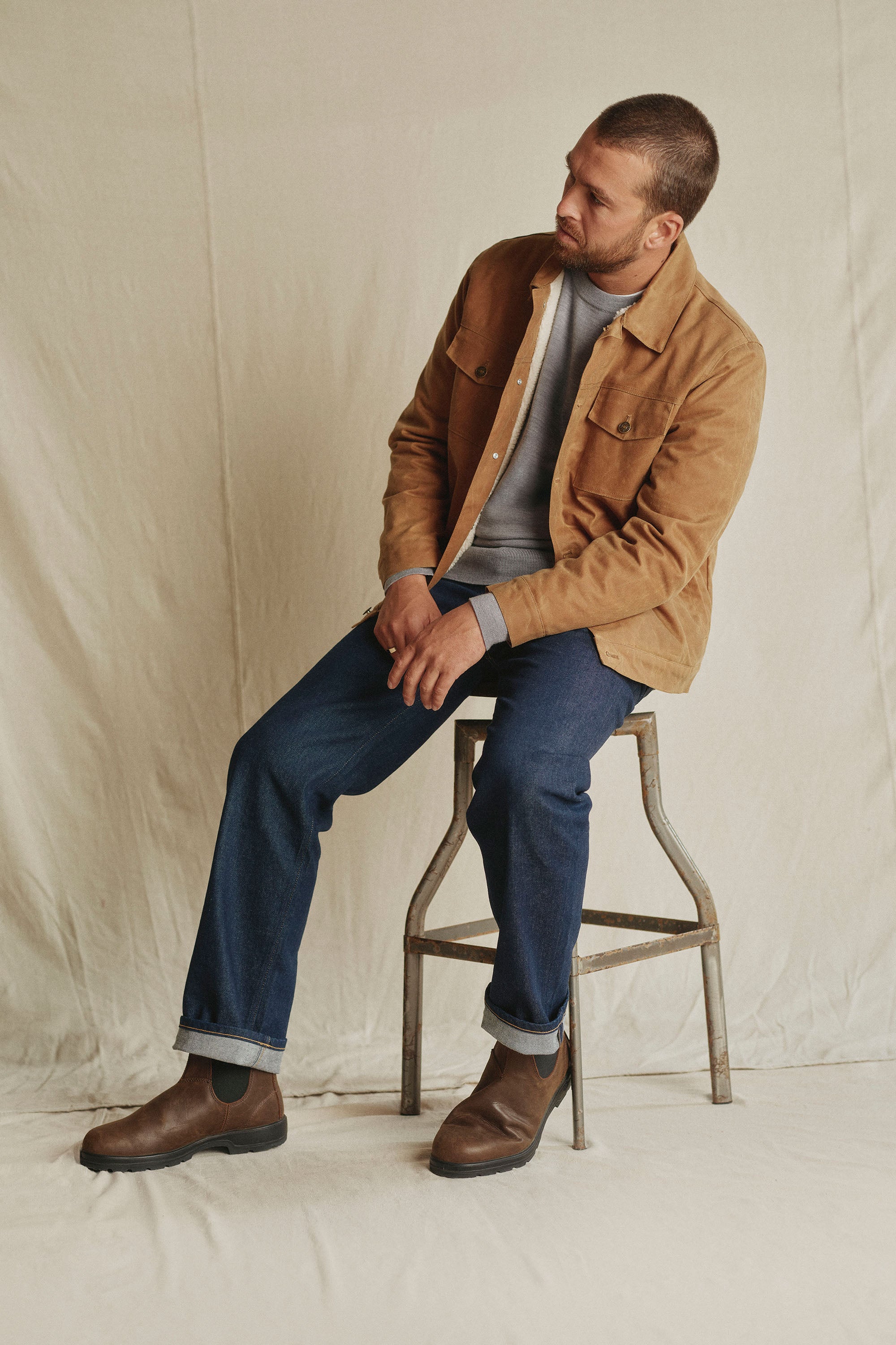 A man with short hair sits on a metal stool against a beige backdrop, wearing a tan jacket over a gray sweater and Barkers Drifter Denim Relax Jean in blue. He looks slightly to the side with brown boots finishing his relaxed look.