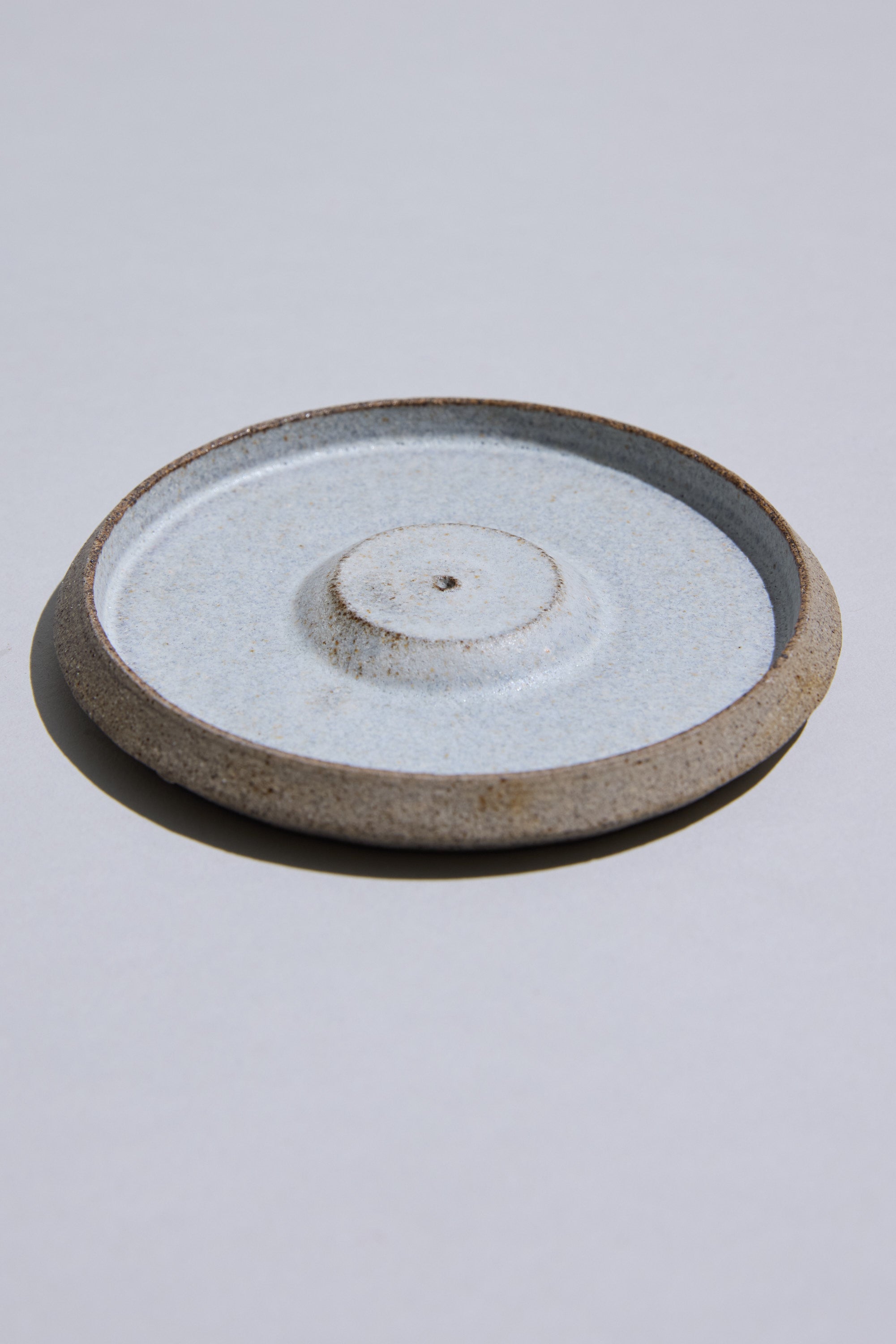 The Studio Foon Incense Dish by Other is a round, shallow New Zealand ceramic plate with a raised center and unglazed rim, featuring a light speckled glaze made from natural clay. It rests on a plain light gray surface.