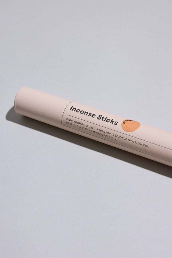A cylindrical beige container labeled EoE Strand Incense Sticks by Earl of East rests on a light gray surface, casting a faint shadow. The label displays instructions and a small orange circle, suggesting fresh mandarin notes inside.