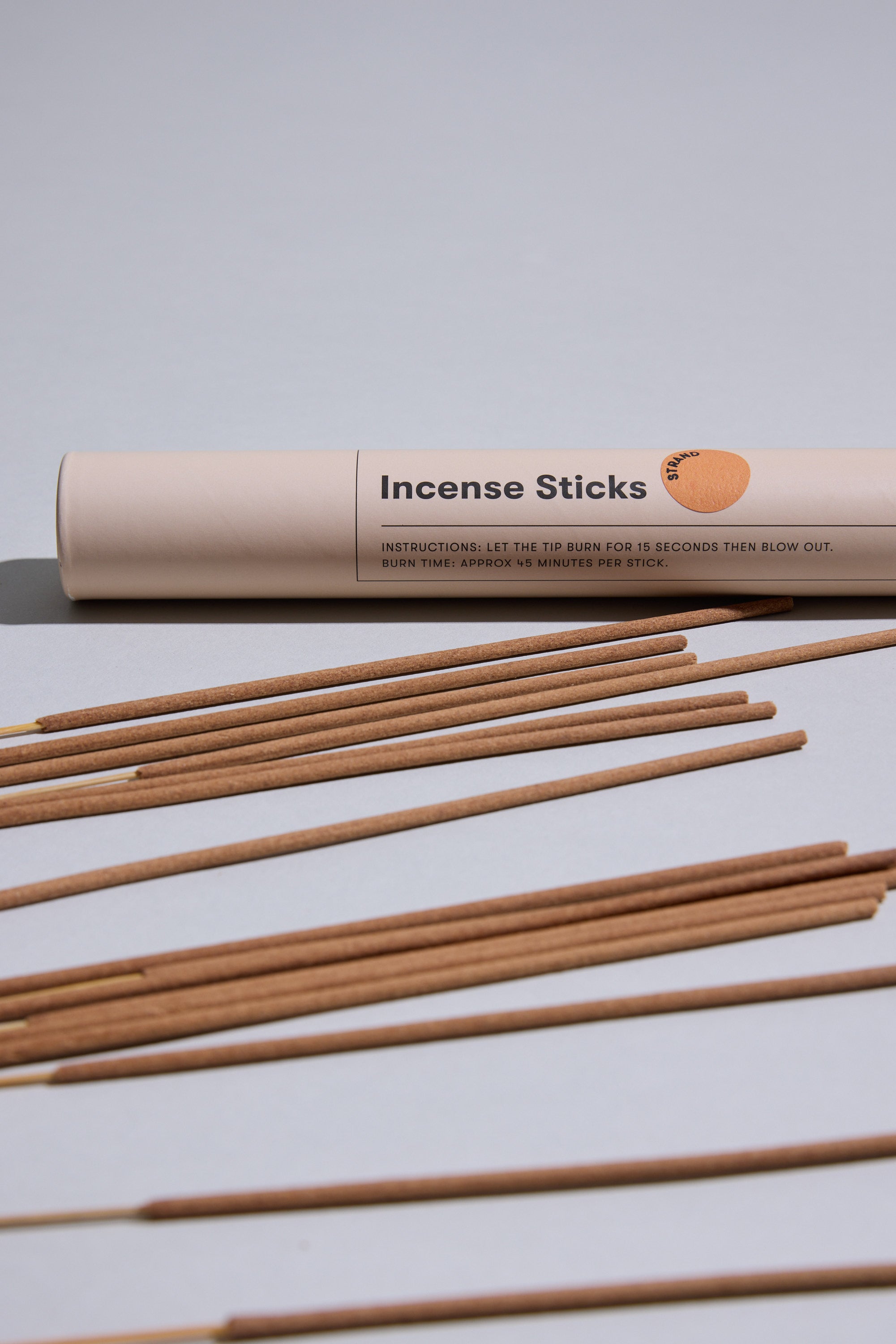 A cylindrical beige Earl of East container labeled "EoE Strand Incense Sticks" rests on a light gray surface, with several birchwood-scented incense sticks arranged in front.