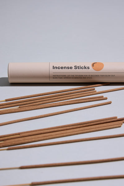 A cylindrical beige Earl of East container labeled "EoE Strand Incense Sticks" rests on a light gray surface, with several birchwood-scented incense sticks arranged in front.