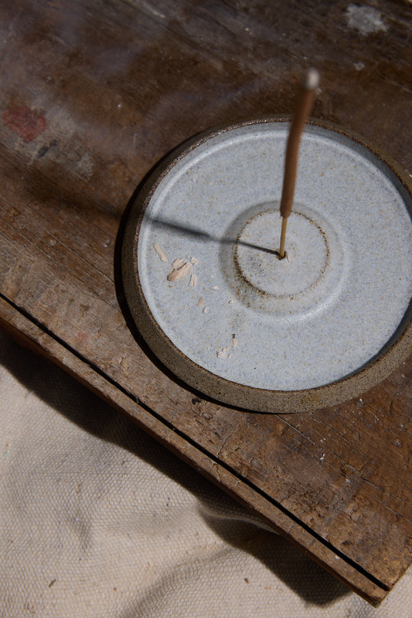 An Earl of East EoE Strand Incense Stick, mandarin-scented, burns on a round ceramic holder atop weathered birchwood. Wisps of smoke rise as ash and debris scatter across the holder and surface.
