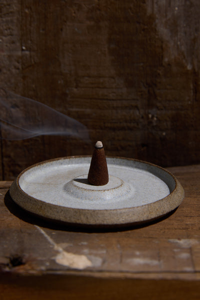 An EoE Smoke And Musk Incense Cone by Earl of East burns on a round ceramic plate, sending a delicate trail of smoke into the air against a rustic wooden background.