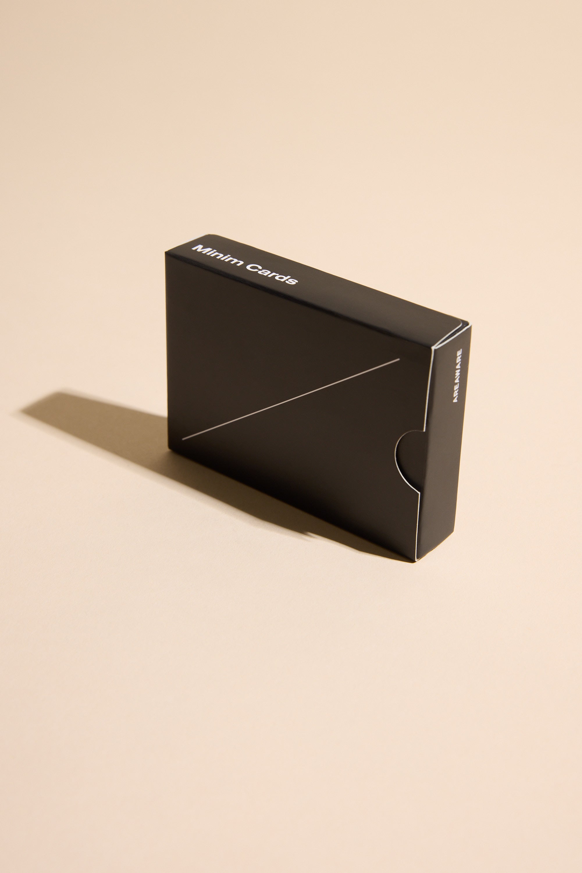 A black rectangular Minim Playing Cards box by Other sits on a beige surface, casting a left shadow. Its minimalistic, geometric-inspired design features a diagonal line and rounded side cutout.