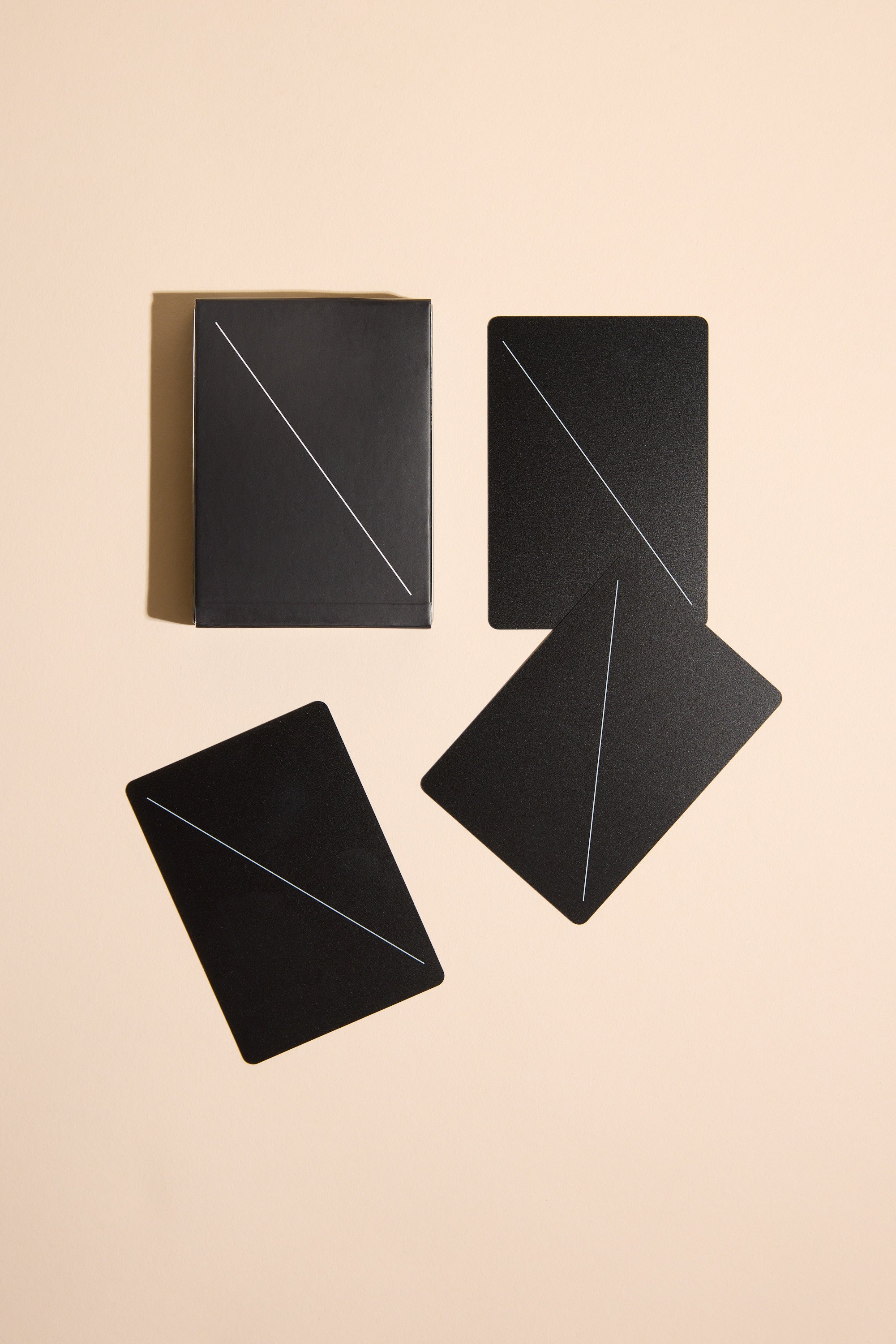 Four Minim Playing Cards by Other, featuring thin diagonal lines, are arranged on a beige surface—three face down and one in a black box.