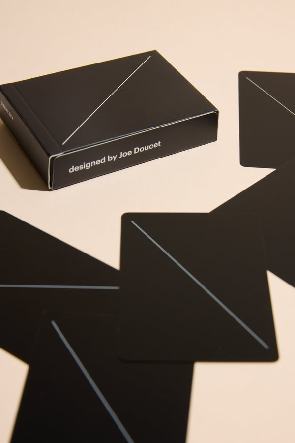 The Minim Playing Cards by Other are minimalist black cards, each with a single diagonal line, scattered beside a matching box labeled "designed by Joe Doucet" on a light surface.