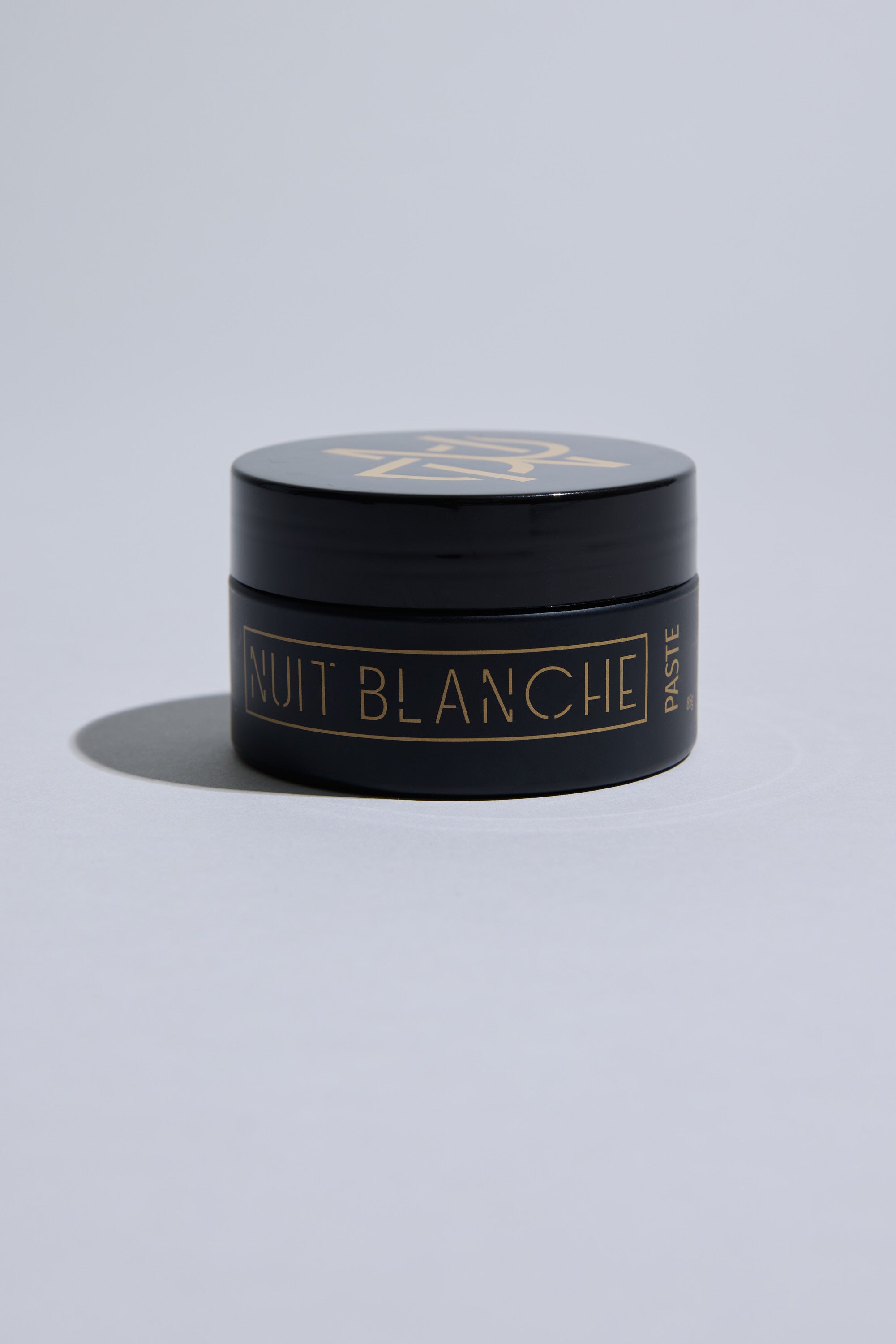 A small black container of Other Nuit Blanche Paste with a gold geometric logo and gold lettering rests on a light gray surface. This medium hold hair paste delivers a natural finish, while its sleek packaging casts a soft shadow.