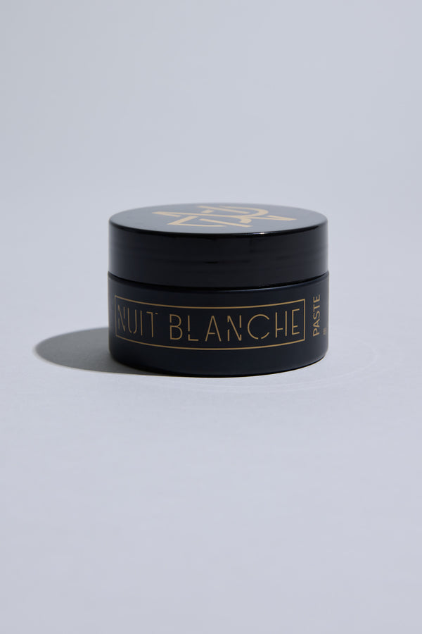 A small black container of Other Nuit Blanche Paste with a gold geometric logo and gold lettering rests on a light gray surface. This medium hold hair paste delivers a natural finish, while its sleek packaging casts a soft shadow.