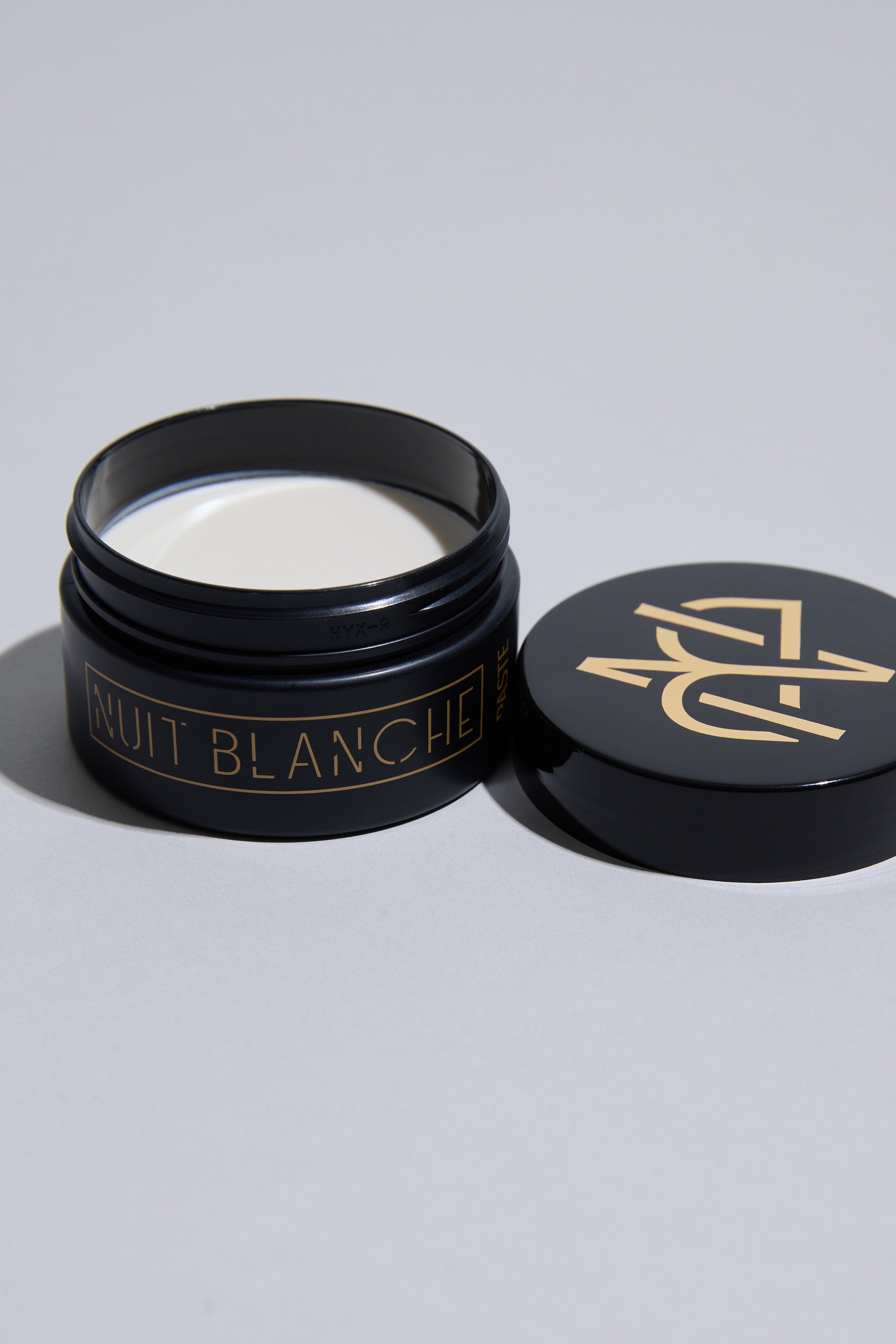 A black jar of Other's Nuit Blanche Paste with gold lettering, filled with white styling paste, sits on a light surface, its matching black and gold lid beside it.