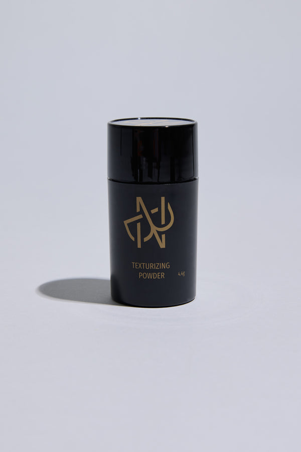 The Nuit Blanche Powder by Other is a black cylindrical container with gold lettering and logo, standing upright on a light gray surface with a subtle shadow—ideal for boosting hair volume and providing a matte finish.