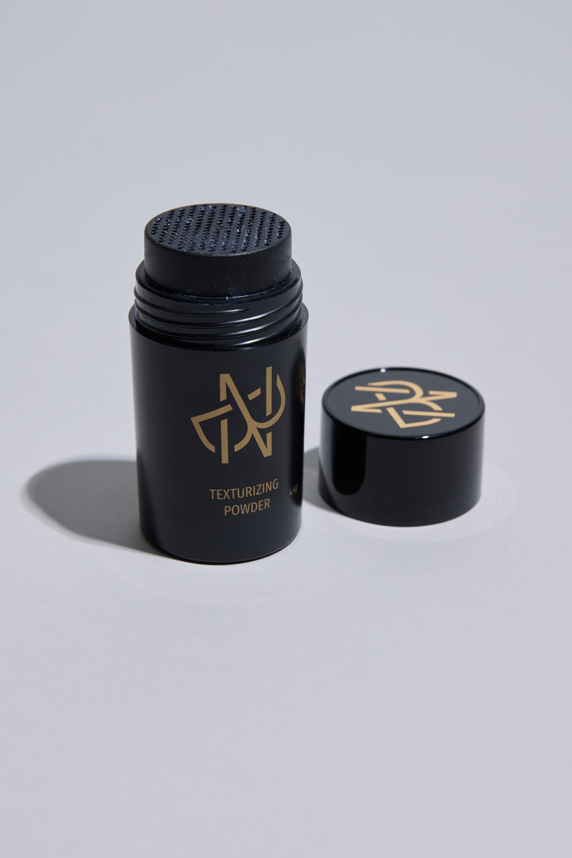 An open black container of Other Nuit Blanche Powder with a gold logo sits on a light gray surface. The cap, also with the gold logo, is to the right. This styling powder boosts hair volume and adds a natural matte finish.