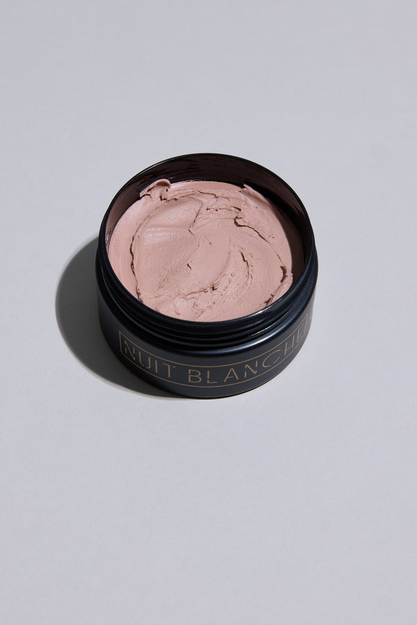The Other Nuit Blanche Clay comes in a black round container with gold text, holding smooth pale pink styling clay. Made in New Zealand, the open container rests on a plain light gray surface.