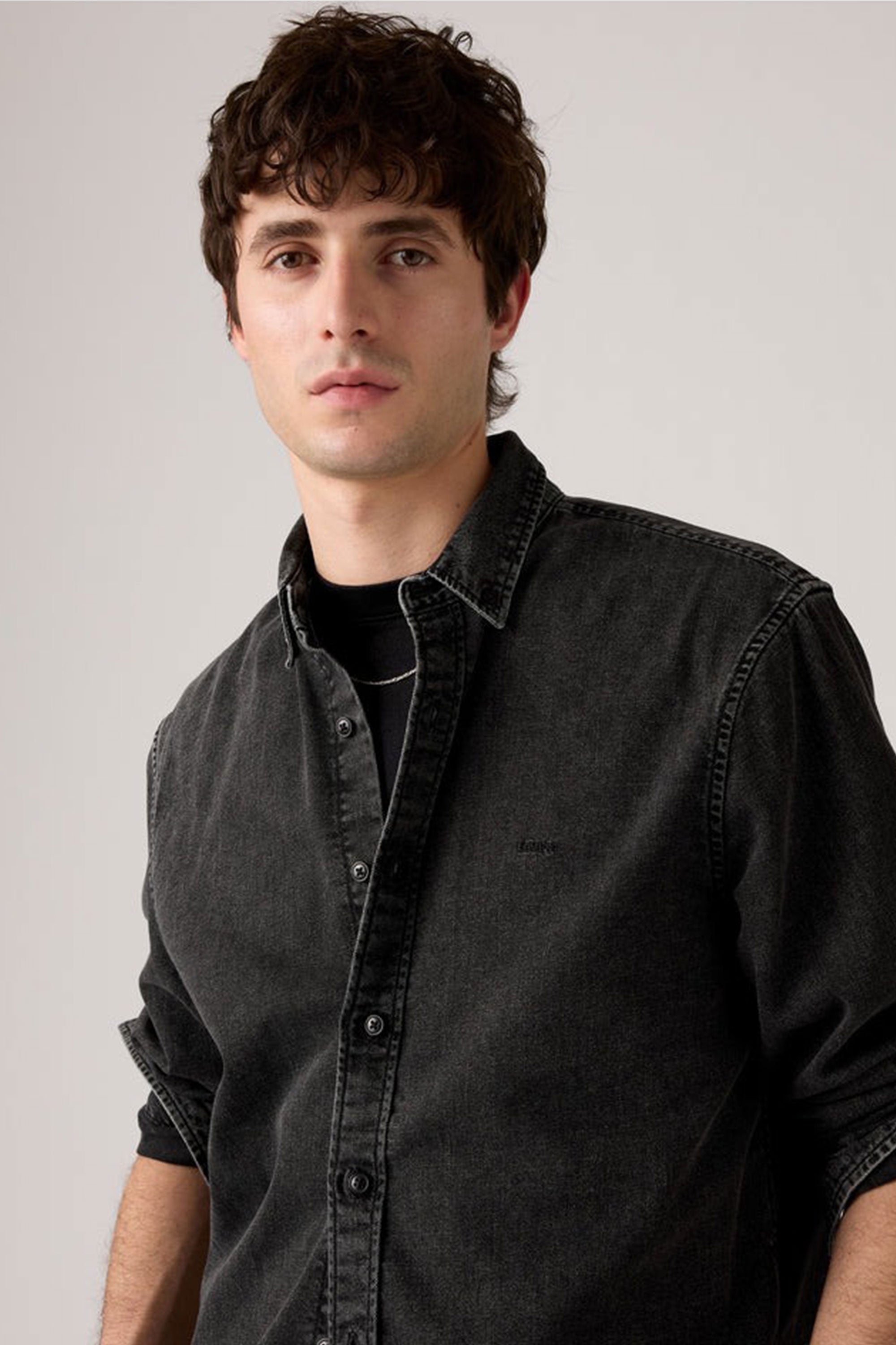 A young man with wavy brown hair wears a Levis Authentic Button Down Long Sleeve Shirt in a relaxed fit over a black t-shirt, standing against a plain light background and looking calmly at the camera.