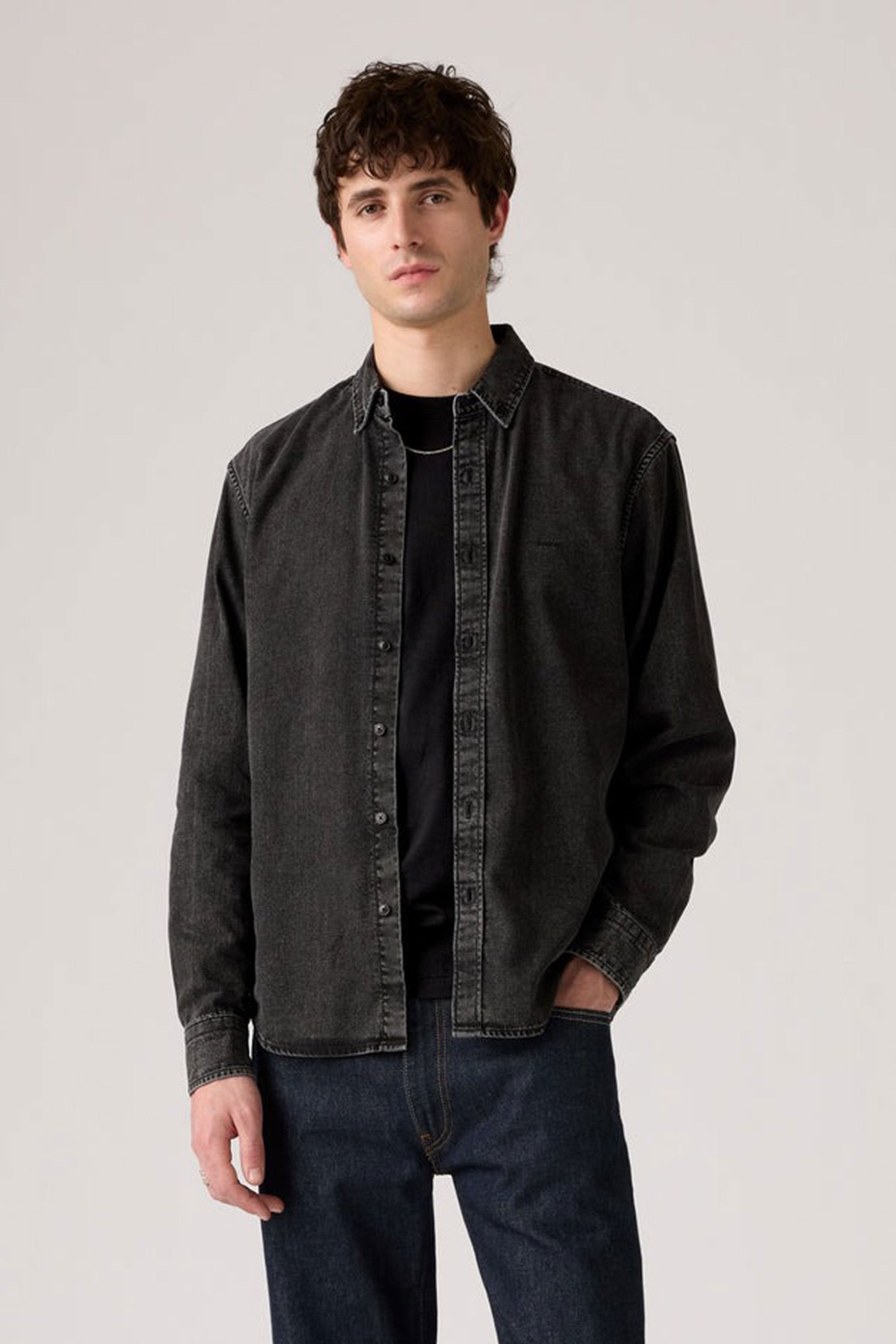 A young man with short, wavy brown hair wears a Levis Authentic Button Down Long Sleeve Shirt by Levis in black denim over a black t-shirt and dark blue jeans, standing against a plain light gray background with a neutral expression.