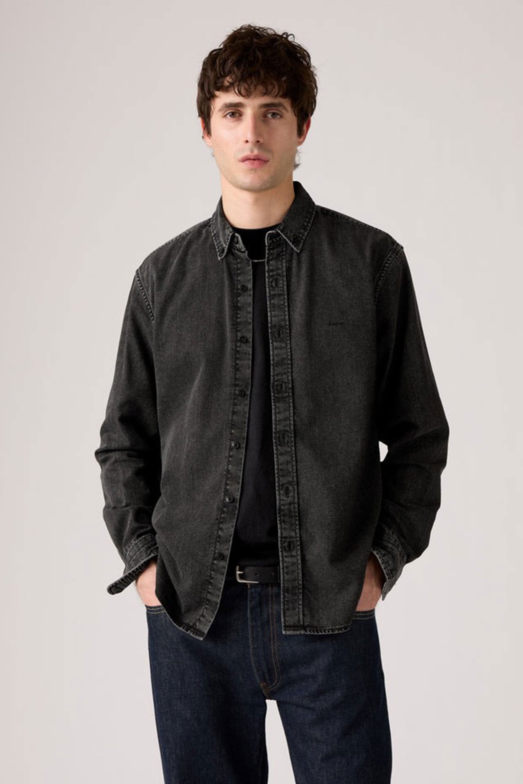 A young man with short, curly brown hair stands against a plain light background, wearing a Levis Authentic Button Down Long Sleeve Shirt over a black t-shirt and dark blue jeans, his hands casually in his pockets.