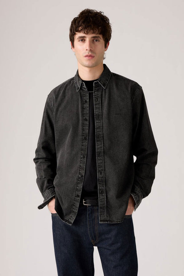 A young man with short, curly brown hair stands against a plain light background, wearing a Levis Authentic Button Down Long Sleeve Shirt over a black t-shirt and dark blue jeans, his hands casually in his pockets.
