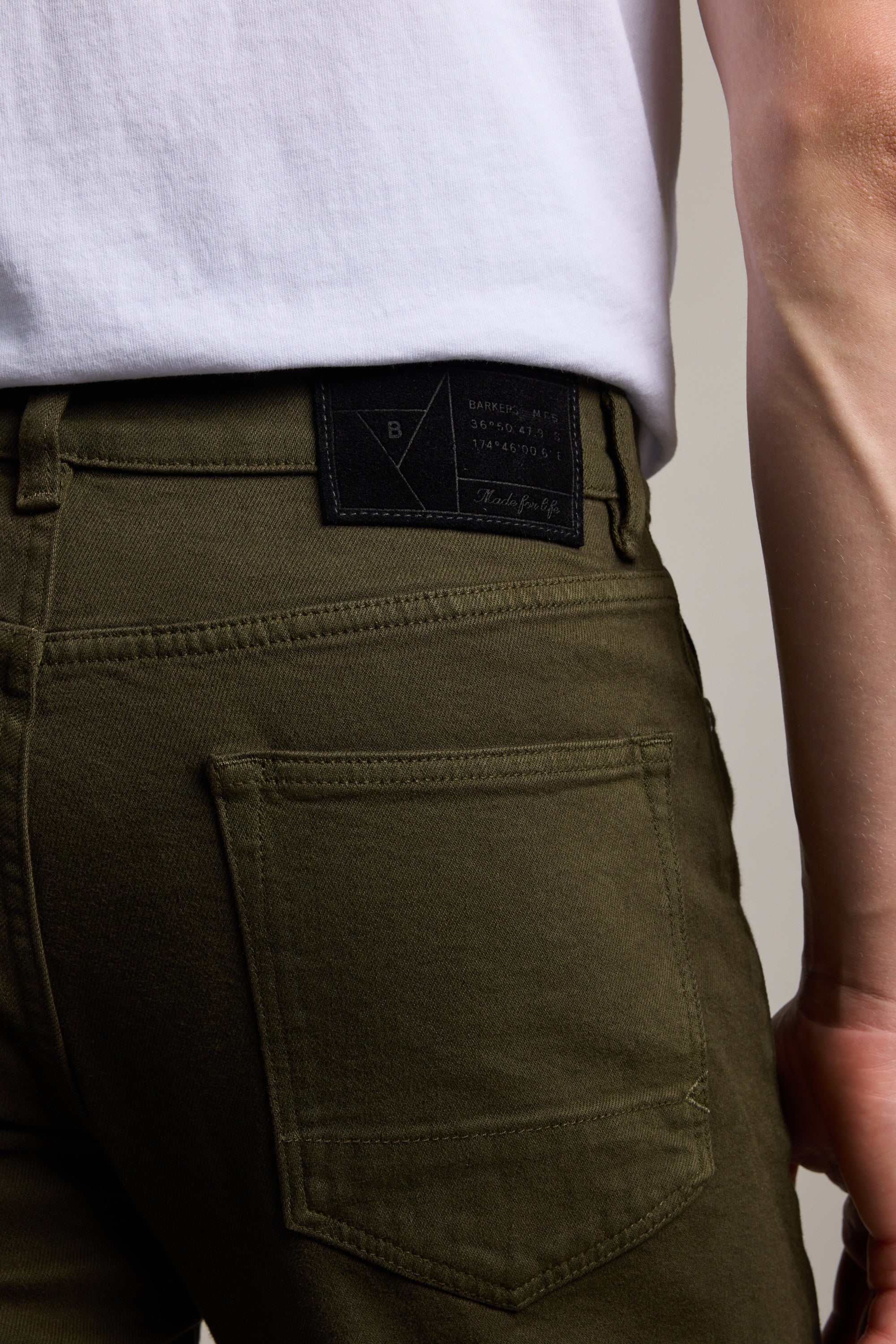 Close-up of a person wearing Barkers’ Wanderer Straight Coloured Jean in olive green with sustainable denim, white shirt, and focus on the back pocket and black denim label near the waistband. The wearer’s arm is partially visible.