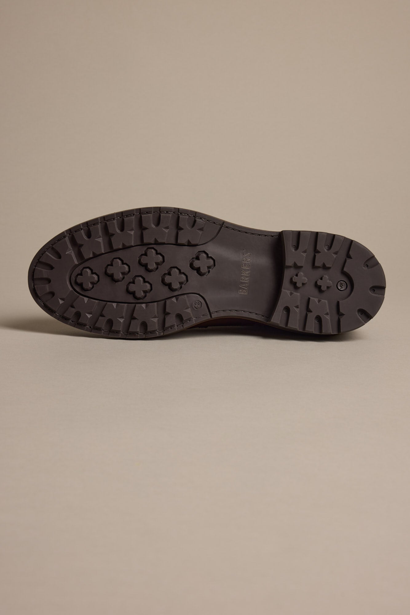 The image displays the sole of the Barkers Carlton Boot, a dark-colored men's luxury shoe with cross-shaped treads, circular grips, and central branding, set against a plain beige background.