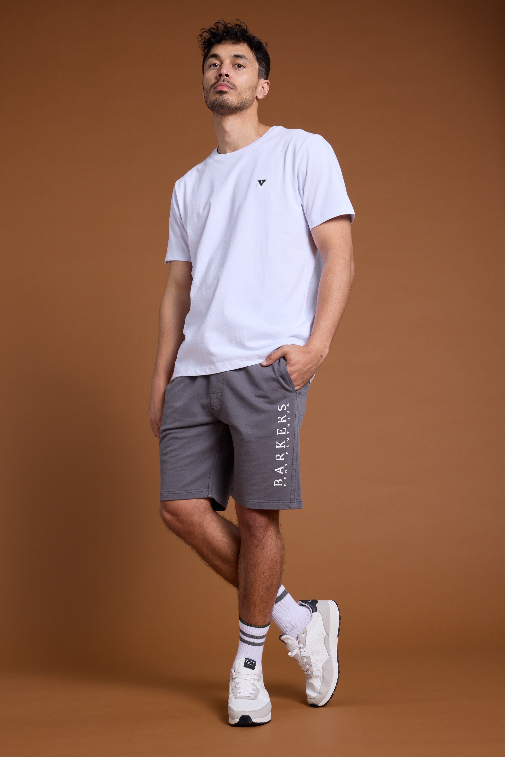 A man with short dark hair stands against a brown background wearing a white T-shirt, Barkers’ Organic Classic Print Track Shorts, white socks, and white sneakers, with one hand in his pocket.