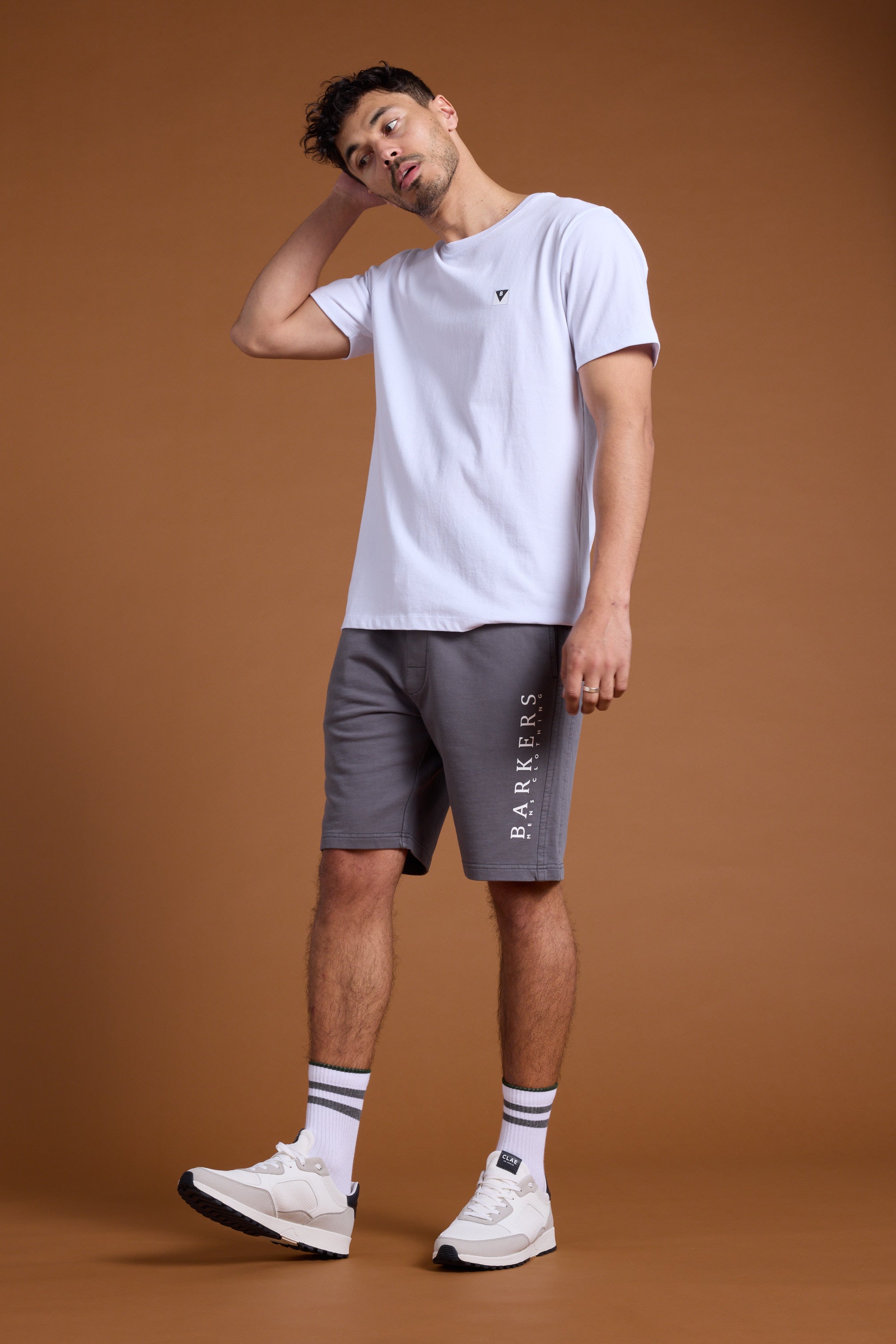 A man stands against a brown background wearing Barkers’ Organic Classic Print Track Short in gray and a white T-shirt. He pairs them with white sneakers and white socks with blue stripes, one hand behind his head, looking slightly to the side.