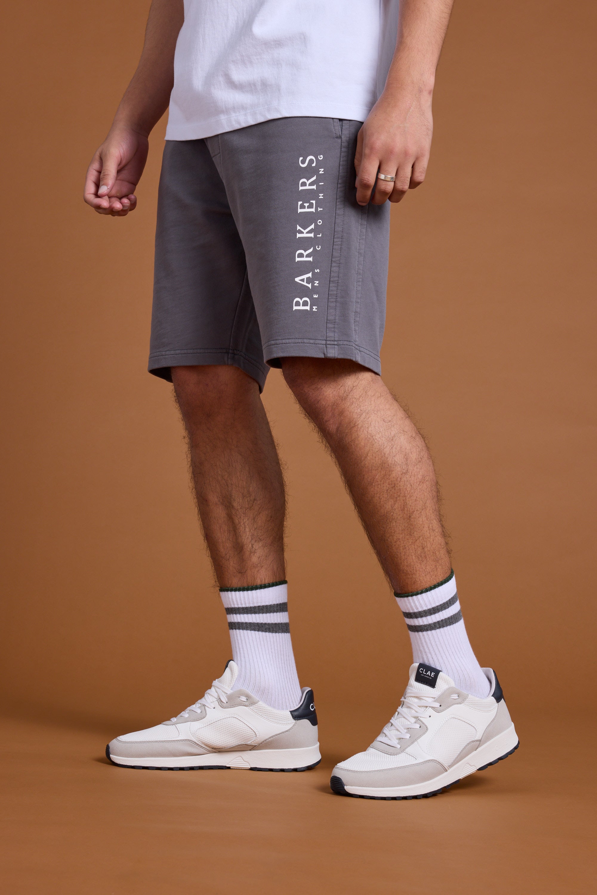 A person models Barkers Organic Classic Print Track Shorts in gray with a white tee, striped white socks, and white sneakers against a plain brown backdrop. Only the lower half is shown.