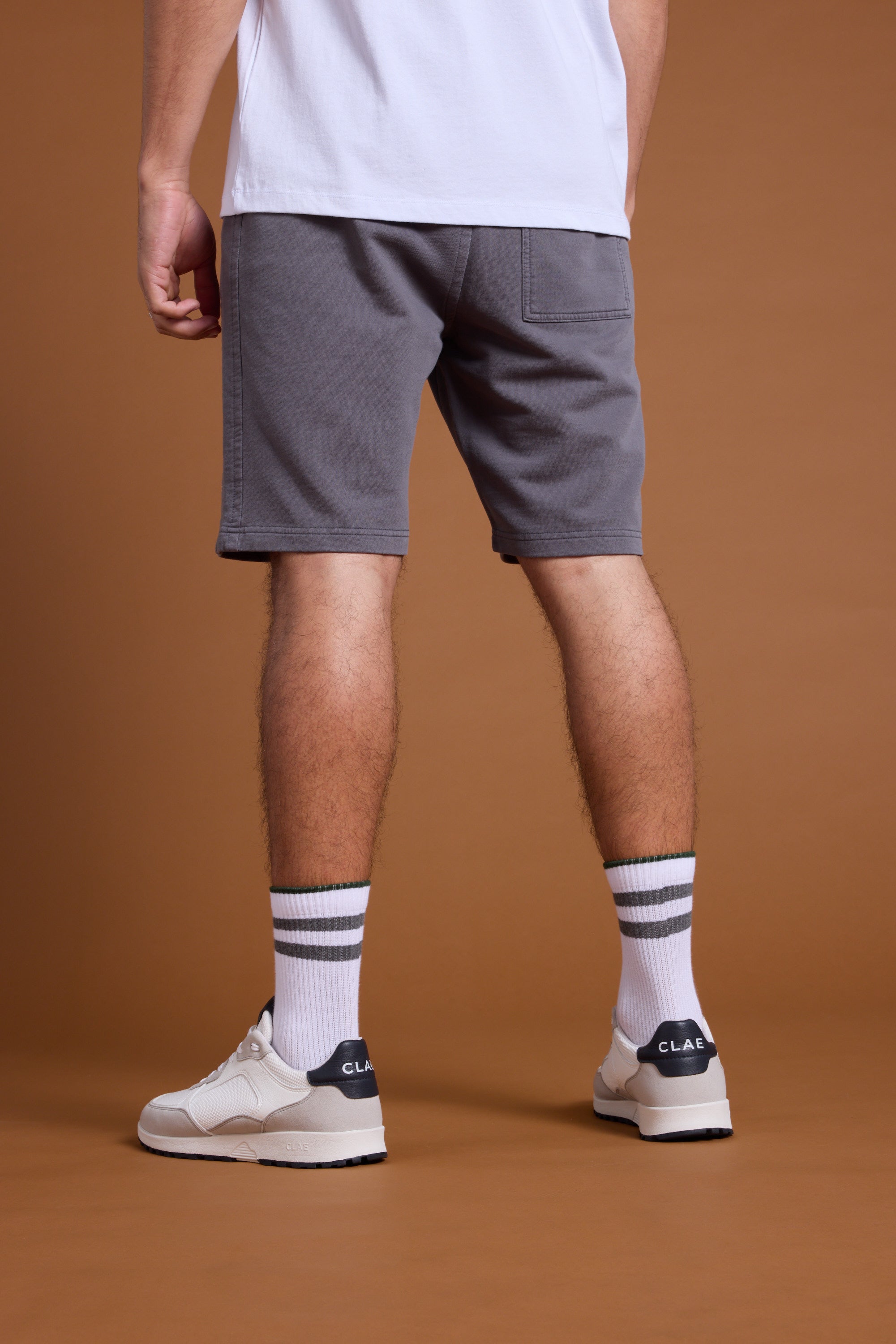 A person stands with their back to the camera, wearing Barkers Organic Classic Print Track Shorts in gray, a white t-shirt, white sneakers, and white socks with gray stripes, set against a brown background.