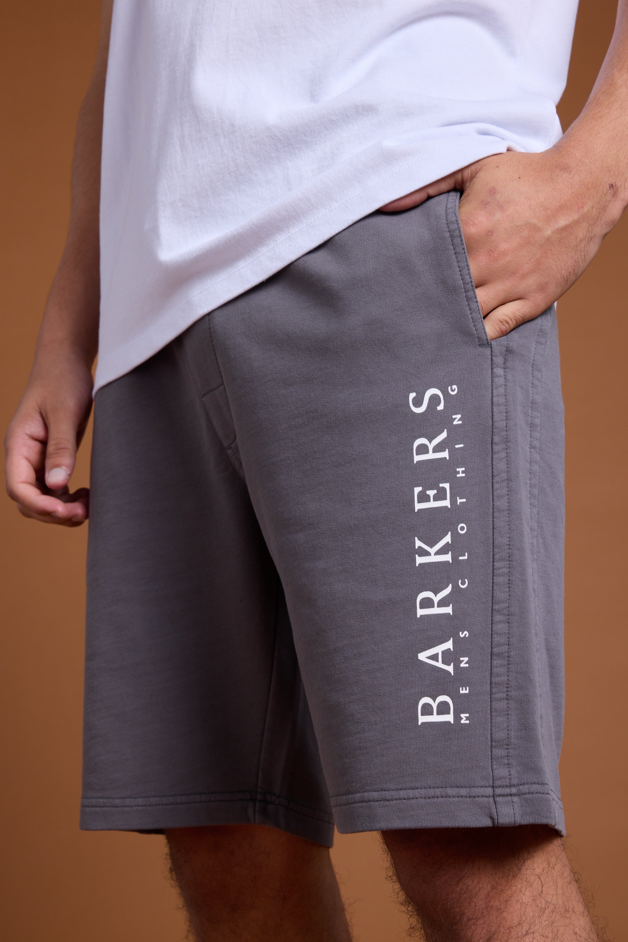 A person wears a white T-shirt and gray Barkers Organic Classic Print Track Short with one hand in a pocket. "BARKERS MENS CLOTHING" is printed vertically on the relaxed fit shorts. The background is brown.