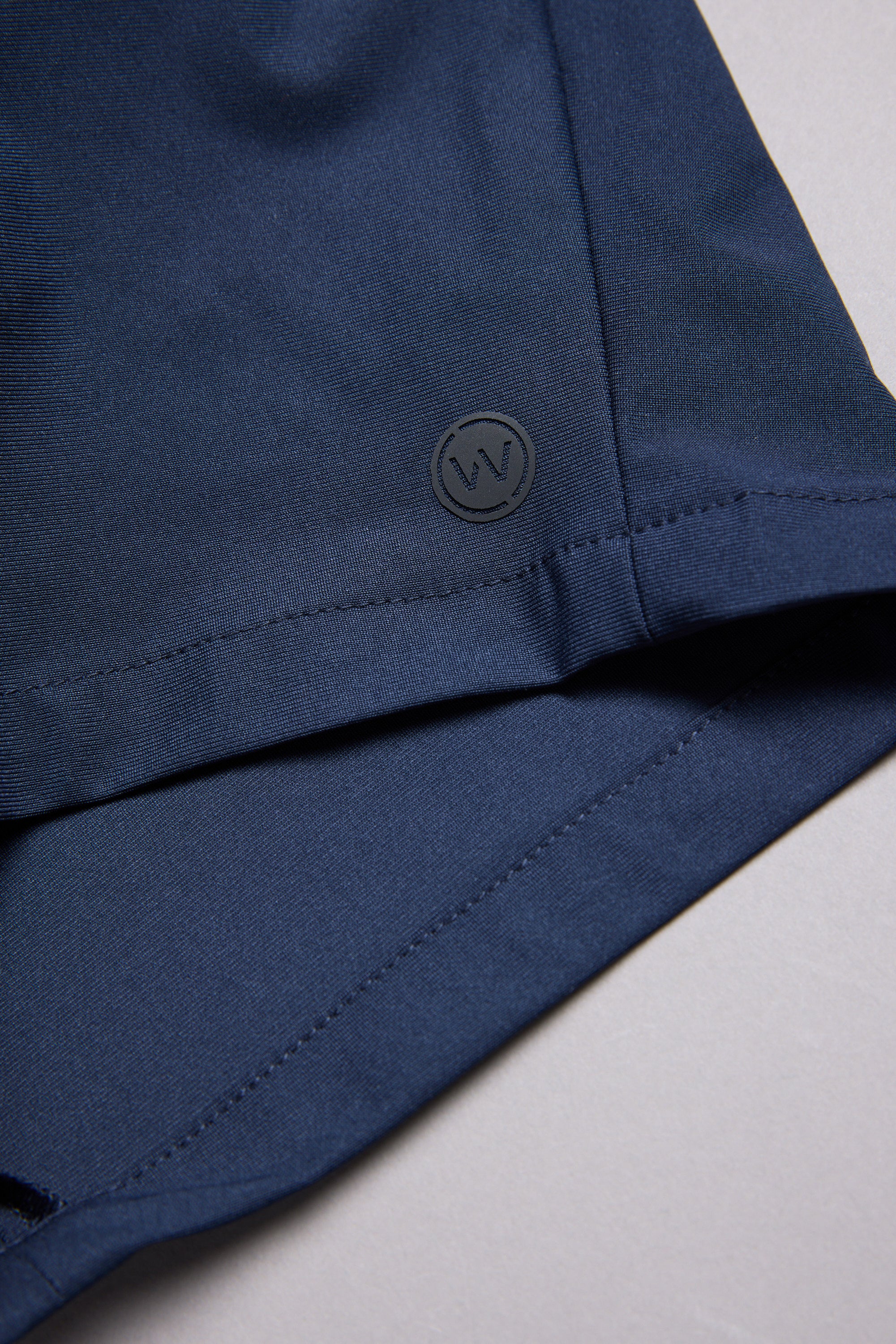 Close-up of the hem of WearEver Switch Short in dark blue, showing a small circular WearEver “W” logo embossed near the seam, set against a light grey background.