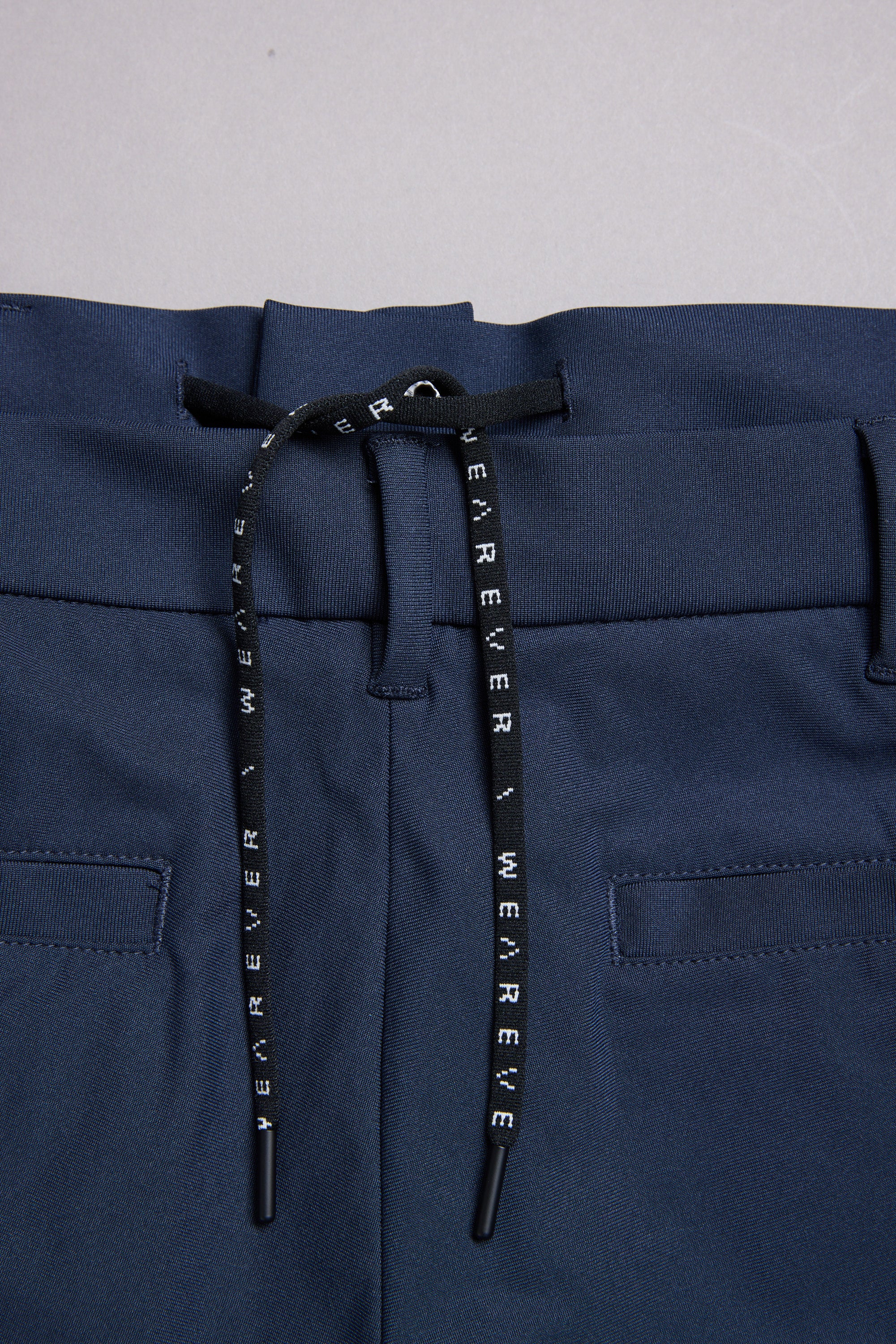 Close-up of the WearEver Long Game Pant: navy performance travel pants with an elastic waistband, black drawstring with white WEAR EVER text, two back welt pockets, and a chino-inspired design.