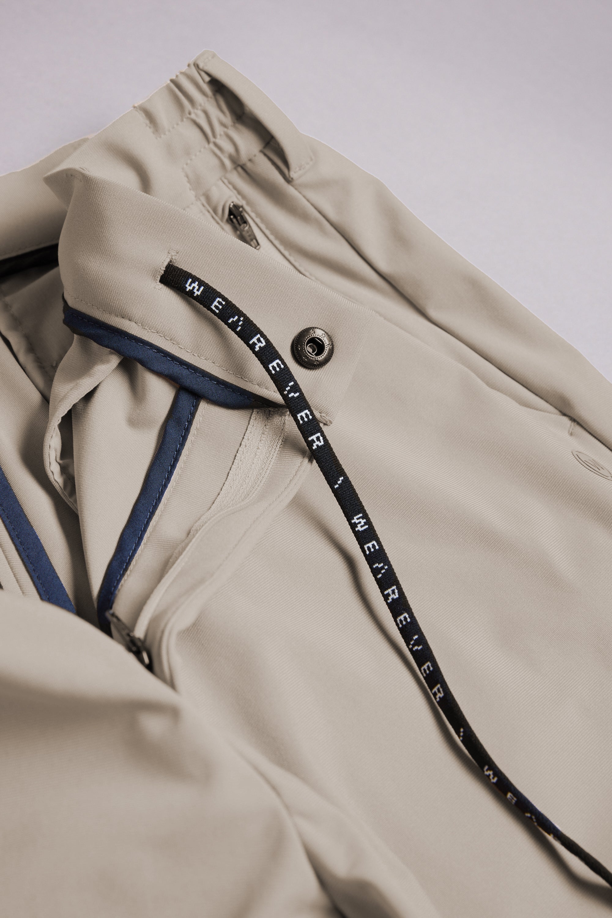A close-up of the WearEver Long Game Pant by WearEver, featuring a beige chino-inspired design with a black drawstring and repeated white WEREVER text, visible button and zipper, plus blue lining—perfect for stylish travel wear.
