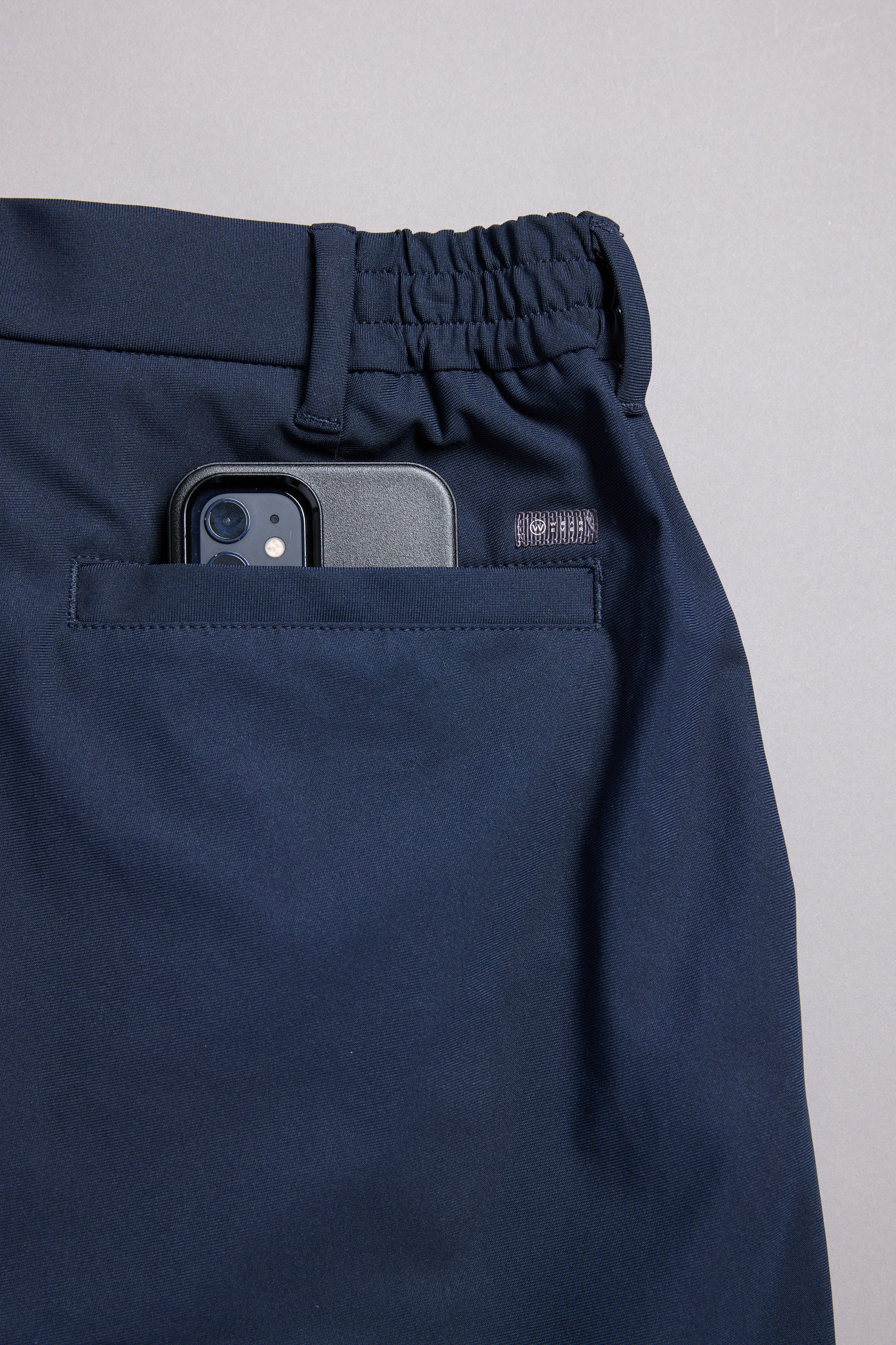 The WearEver Long Game Pant by WearEver is a navy blue, lightweight performance pant with an elastic waistband and a back pocket fitting a black smartphone, featuring chino-inspired design and shown on a light grey background.
