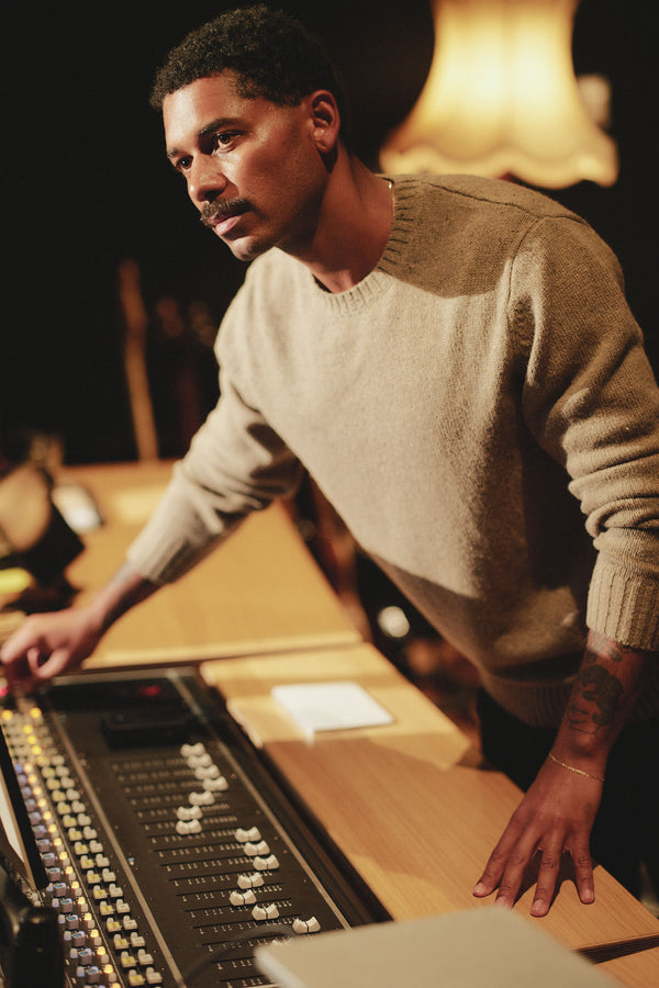 A man wearing the Barkers Anglers Merino Crew stands at a mixing console in a dimly lit recording studio, adjusting sound controls with focus as a warm lamp glows in the background.
